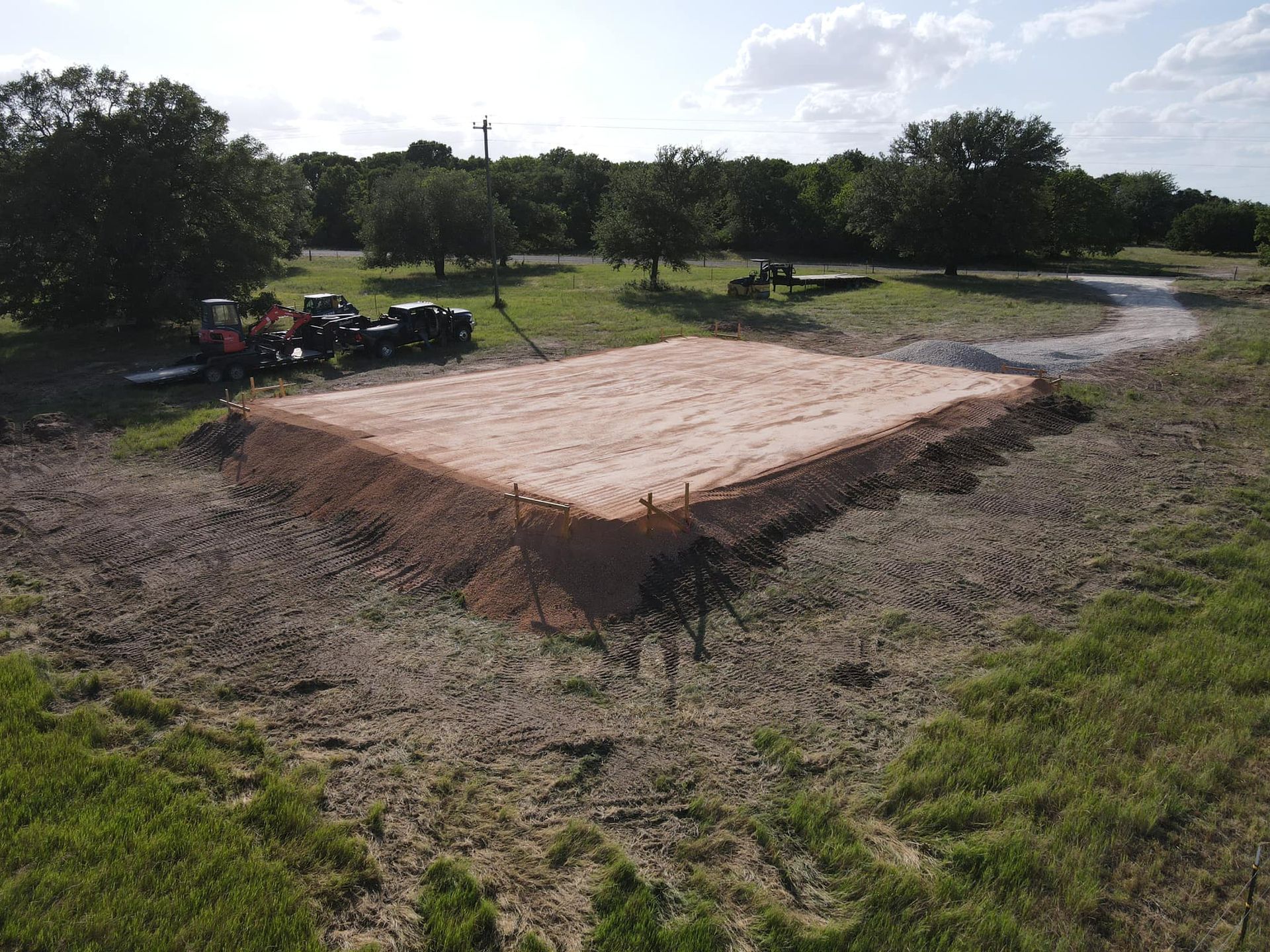 A rectangular earthen platform in a grassy field with trees, construction equipment visible.