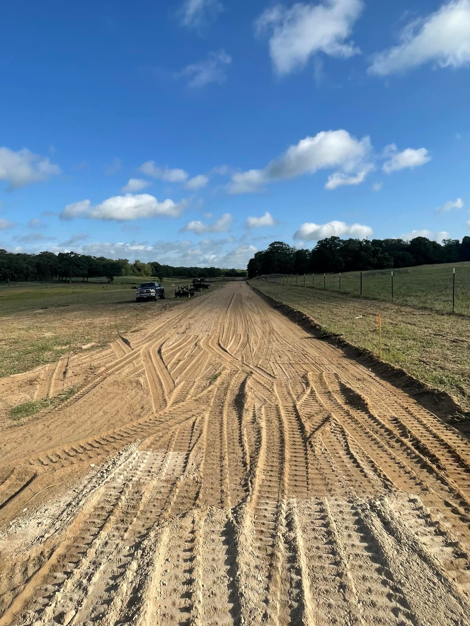 Dirt road under a blue sky with tire tracks, trees, and vehicles in the distance.
