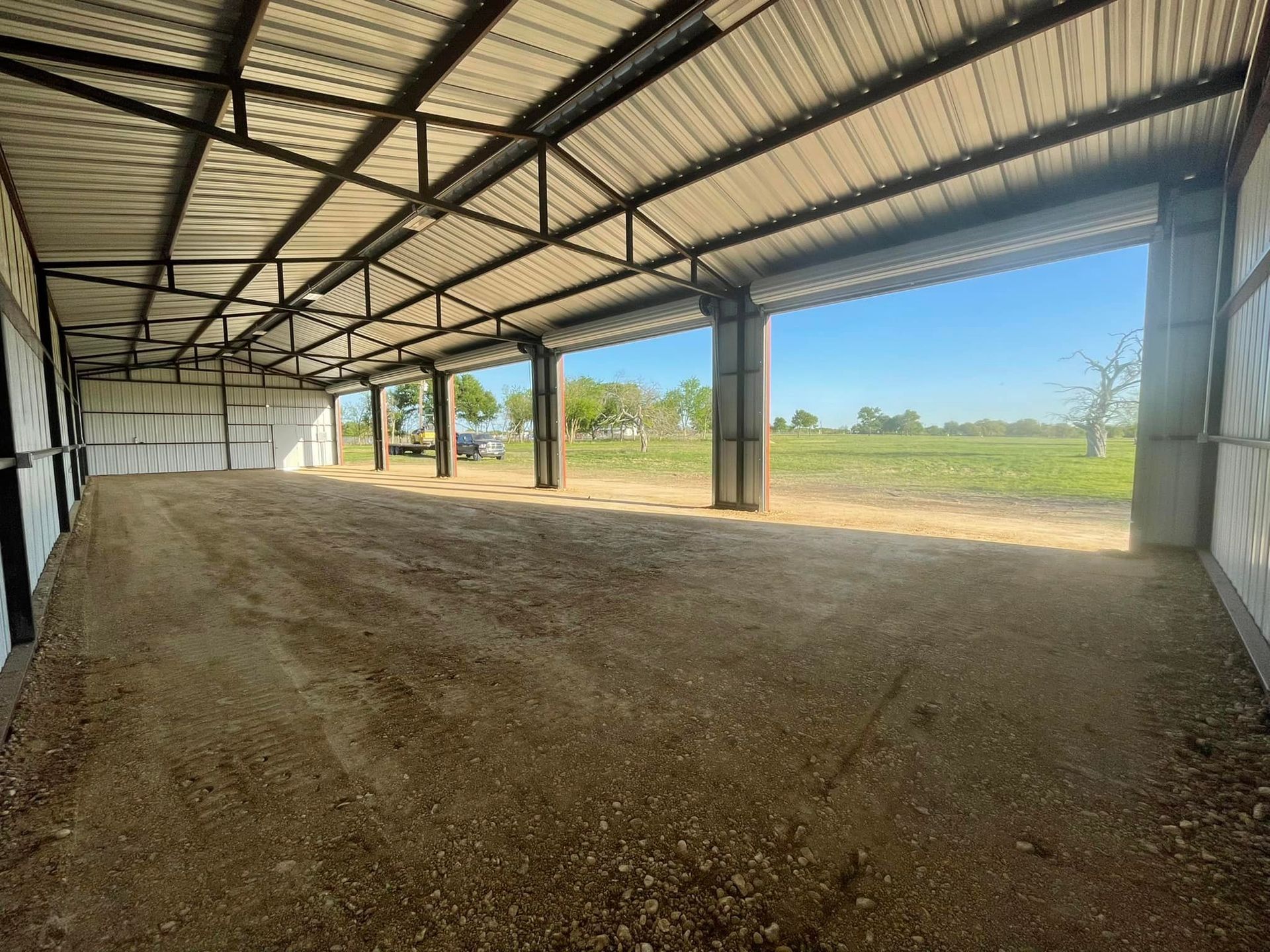 Interior of a metal building with an open front facing a field under a blue sky. The floor is gravel.