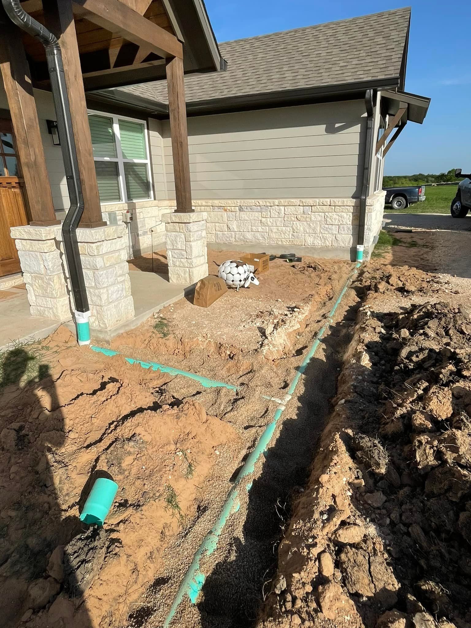 Construction site with trenches and green pipes in front of a house; gravel, dirt, and plumbing.
