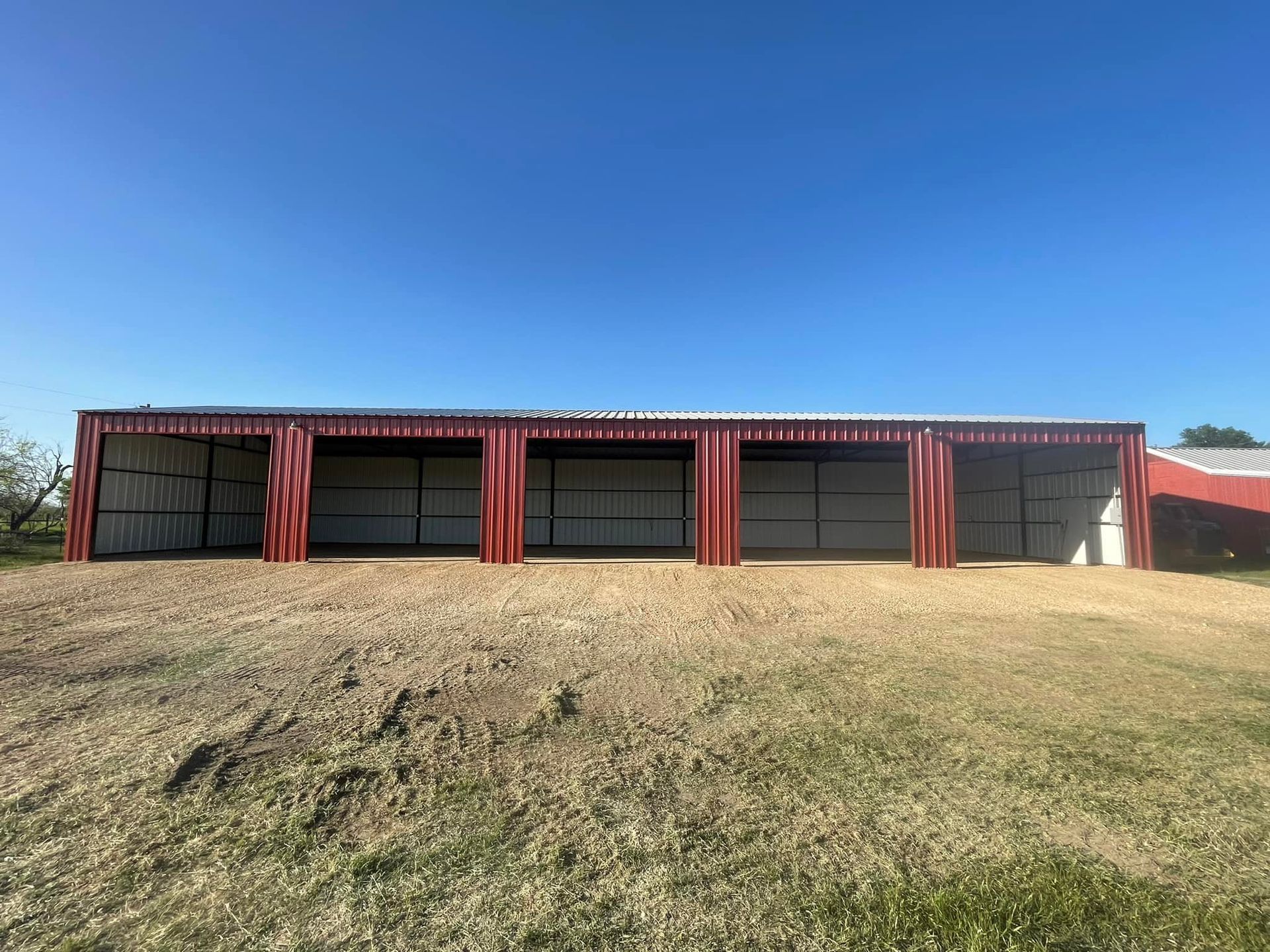 Red metal shed with three open bays on a gravel base, under a blue sky.