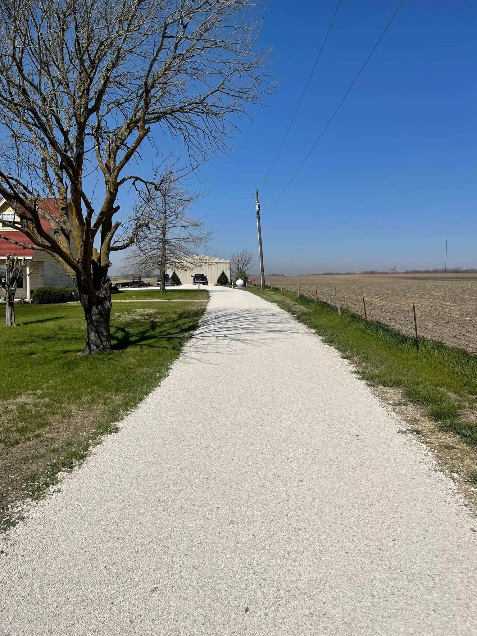A gravel driveway leads to a white building under a blue sky, with a tree on the left and fields on the right.