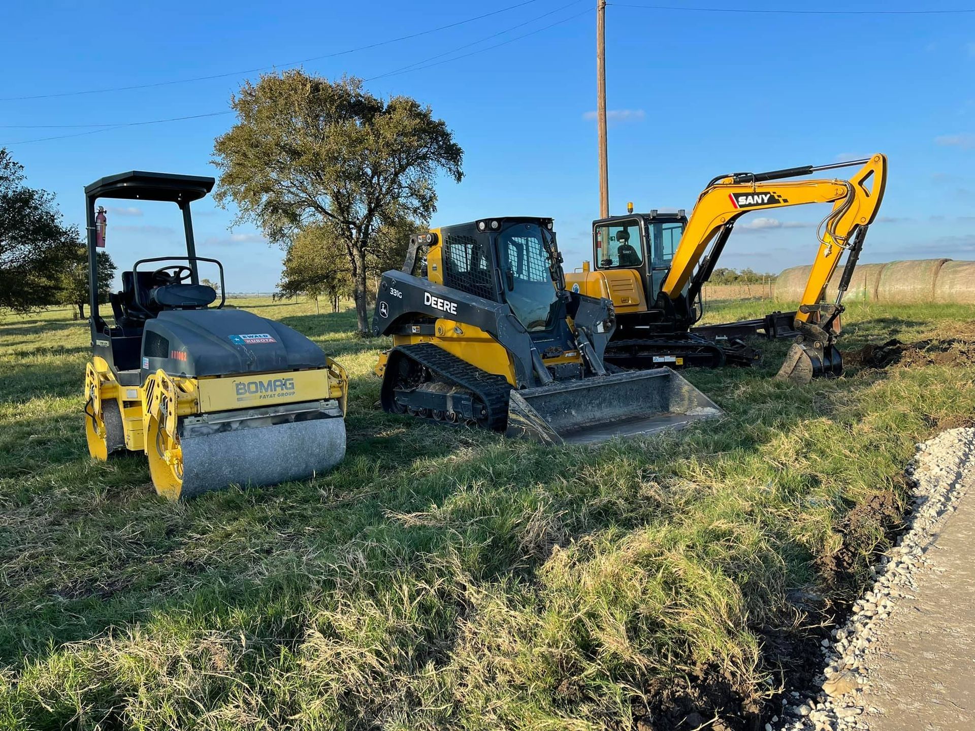 Three pieces of construction equipment on grass: a roller, skid steer, and excavator.