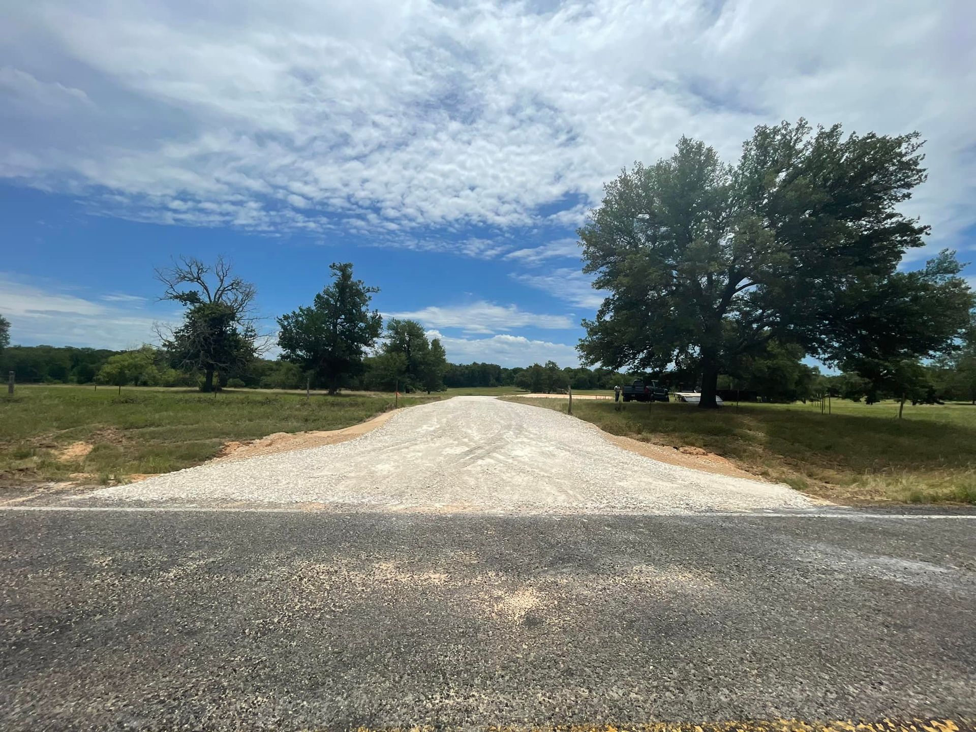 A gravel driveway leads from an asphalt road into a field with trees under a cloudy sky.