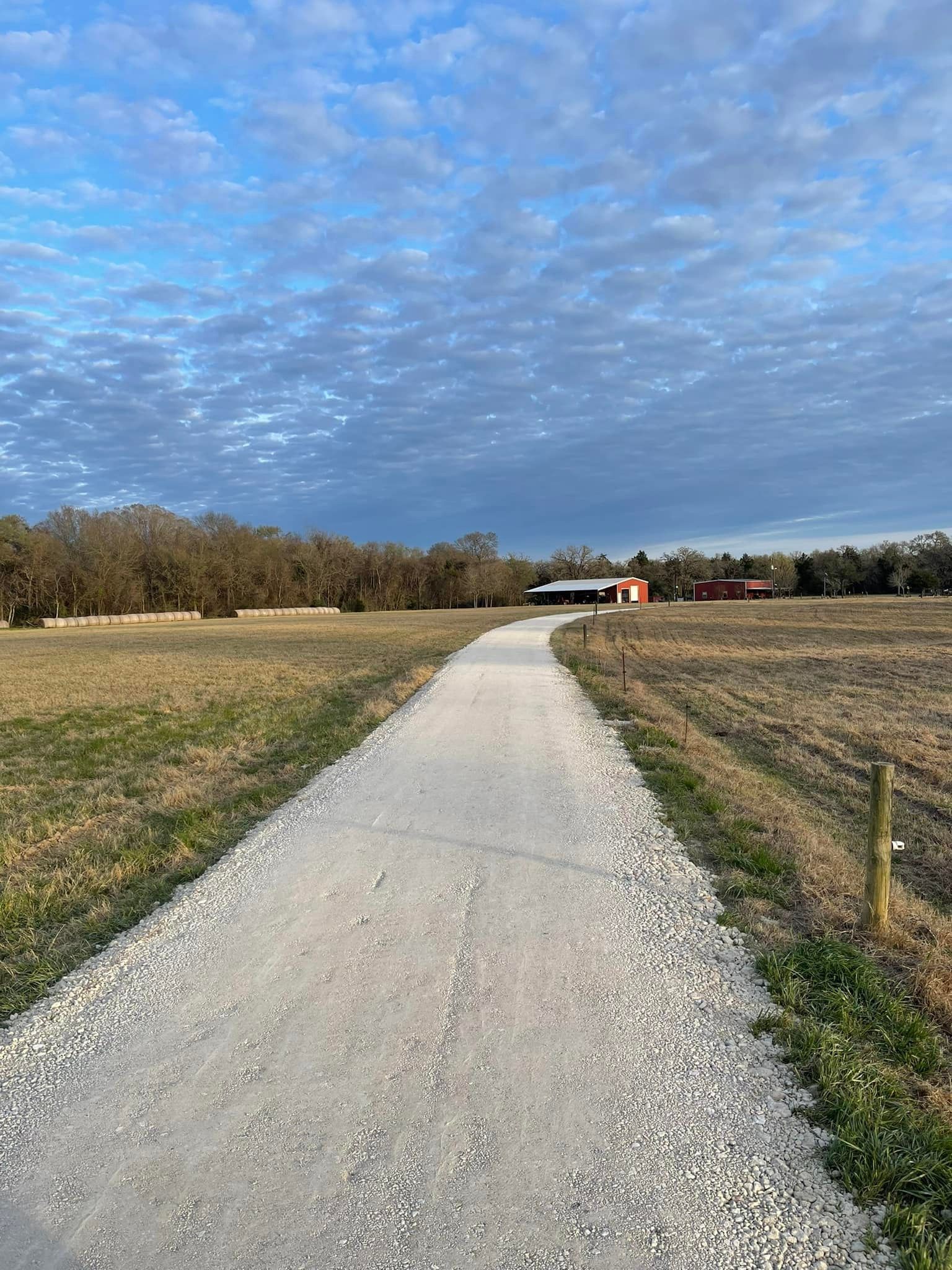 Gravel path leading to red buildings under a cloudy blue sky, flanked by fields and trees.