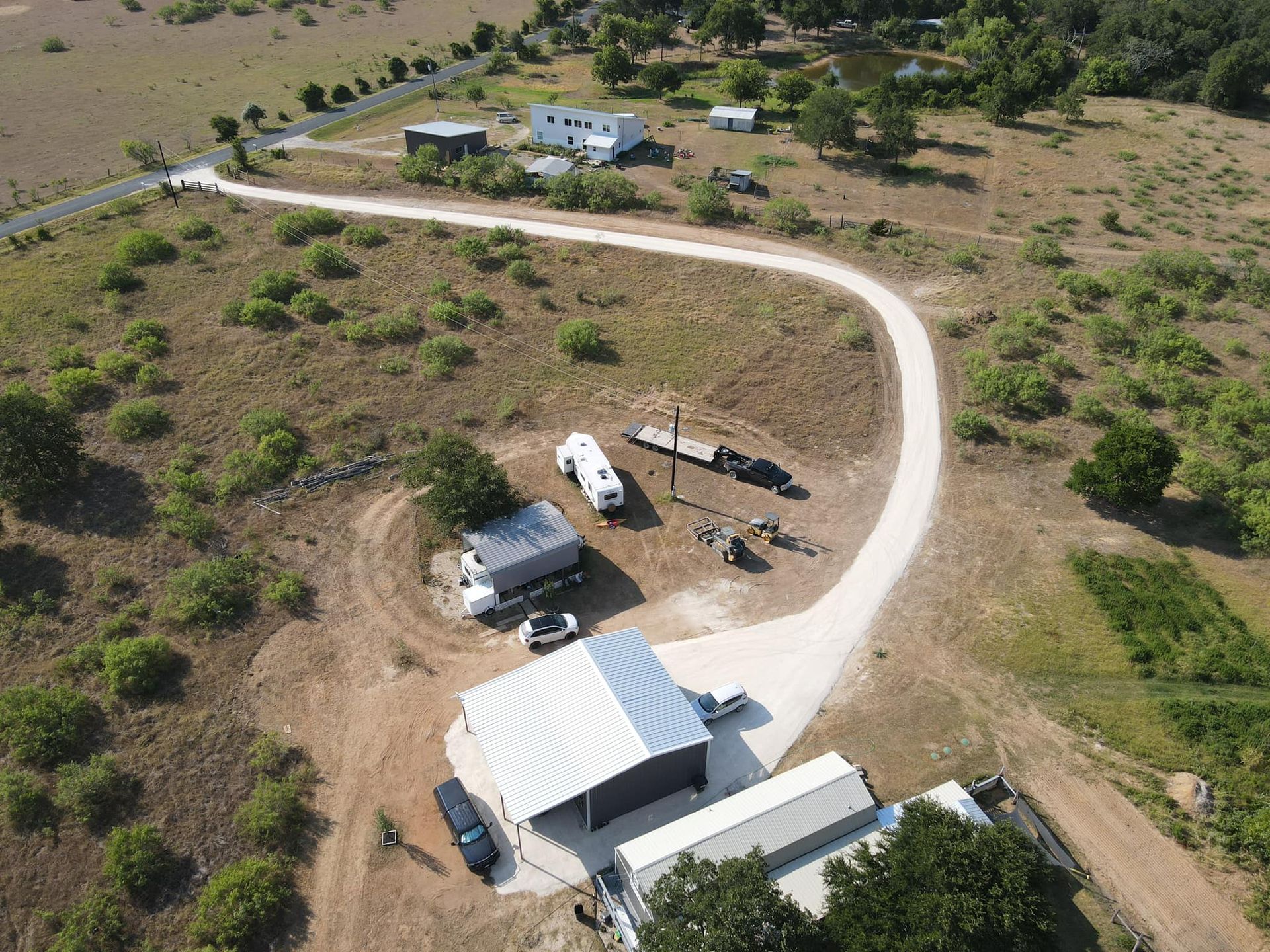 Aerial view: winding gravel road leading to a rural property with buildings, trees, and vehicles on a dry, grassy landscape.