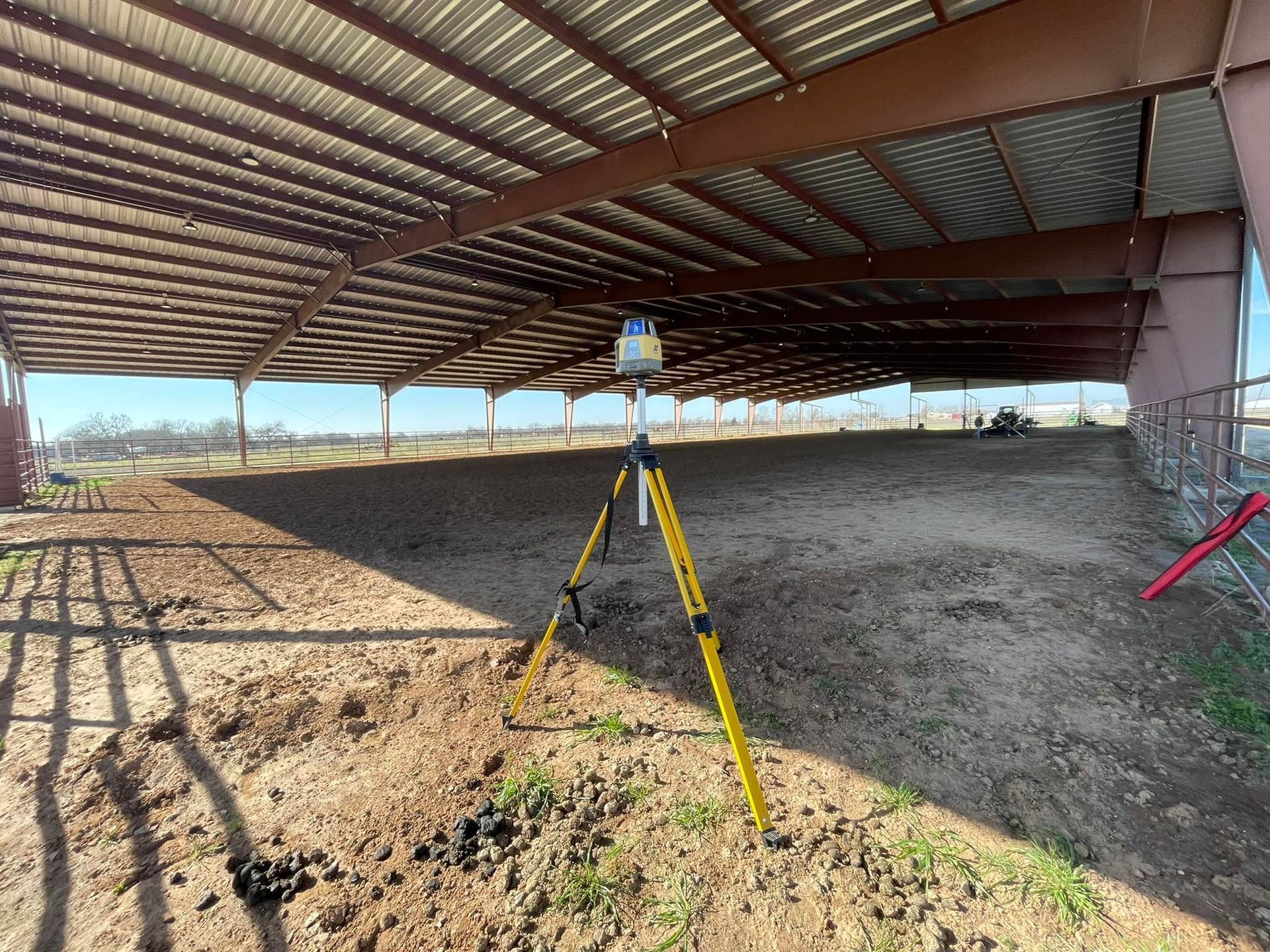A laser scanner on a tripod inside a large, open-air metal-framed building, scanning the ground.