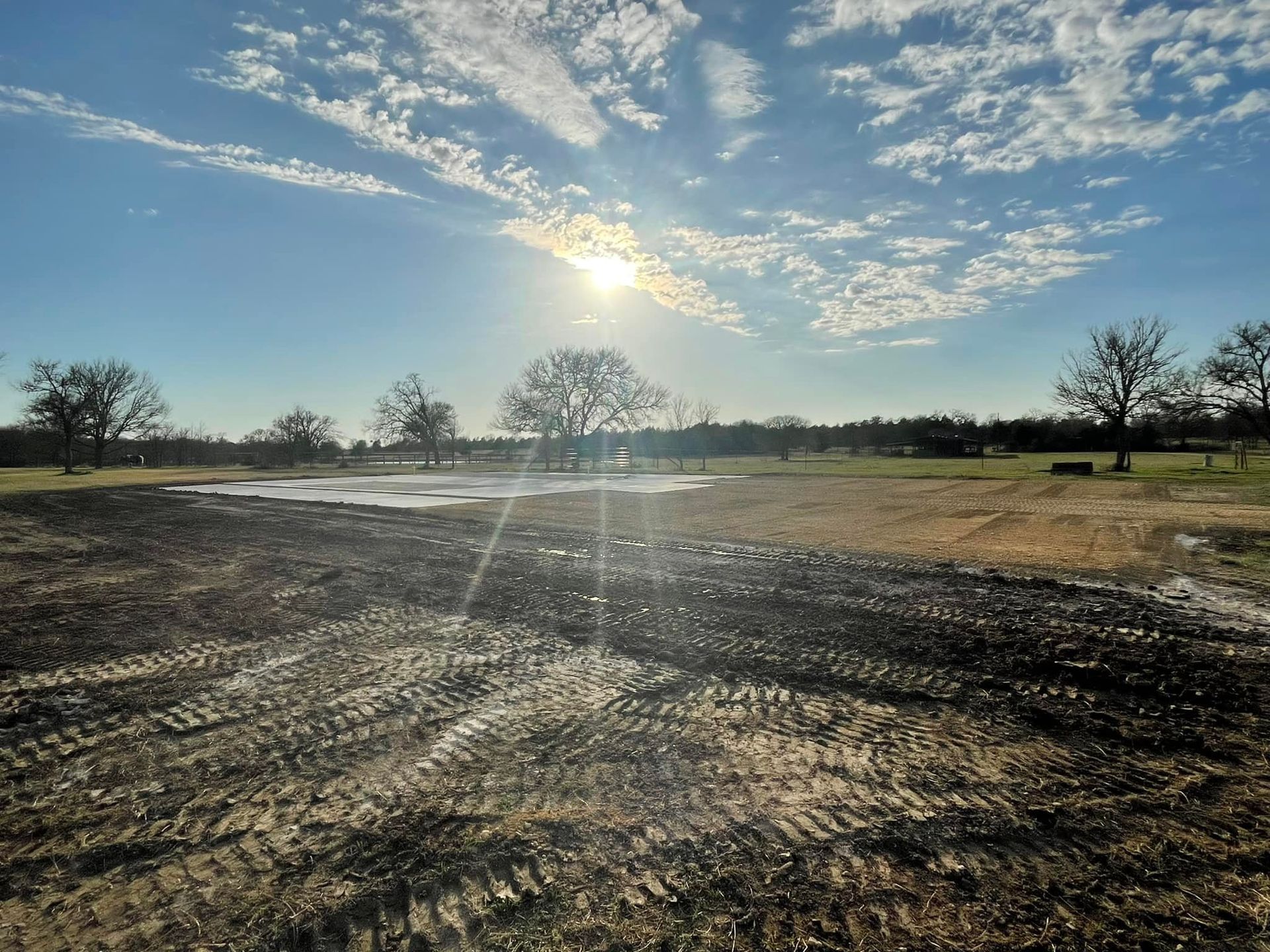 Sunny field with clouds, trees, and muddy ground with tire tracks.