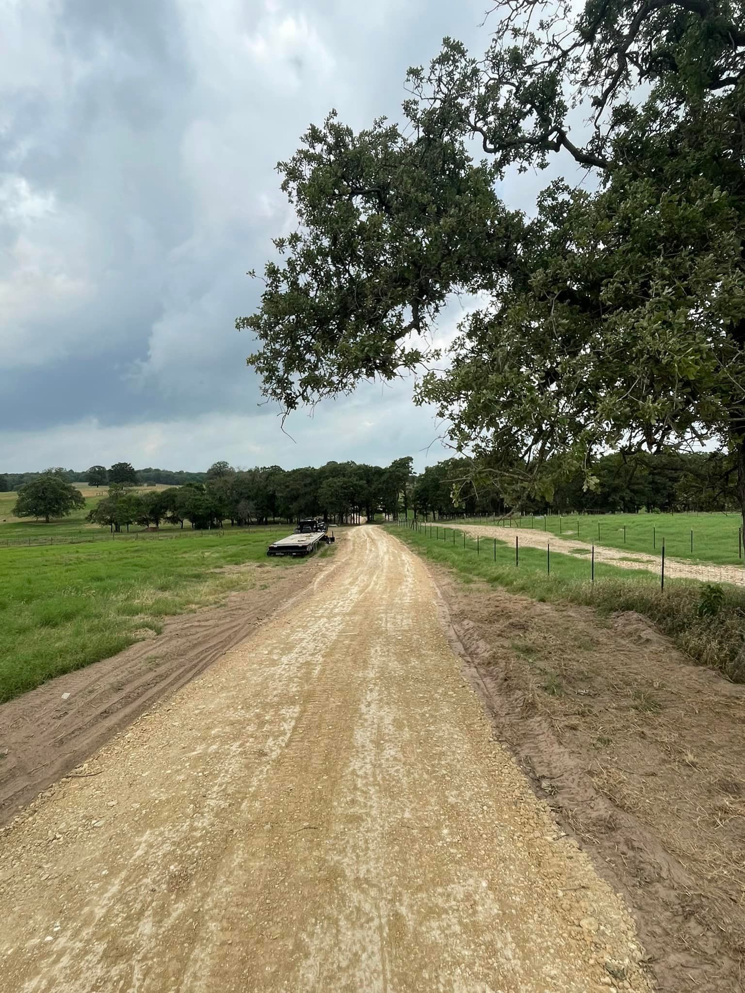 Gravel road leads through a green field toward distant trees under a cloudy sky.