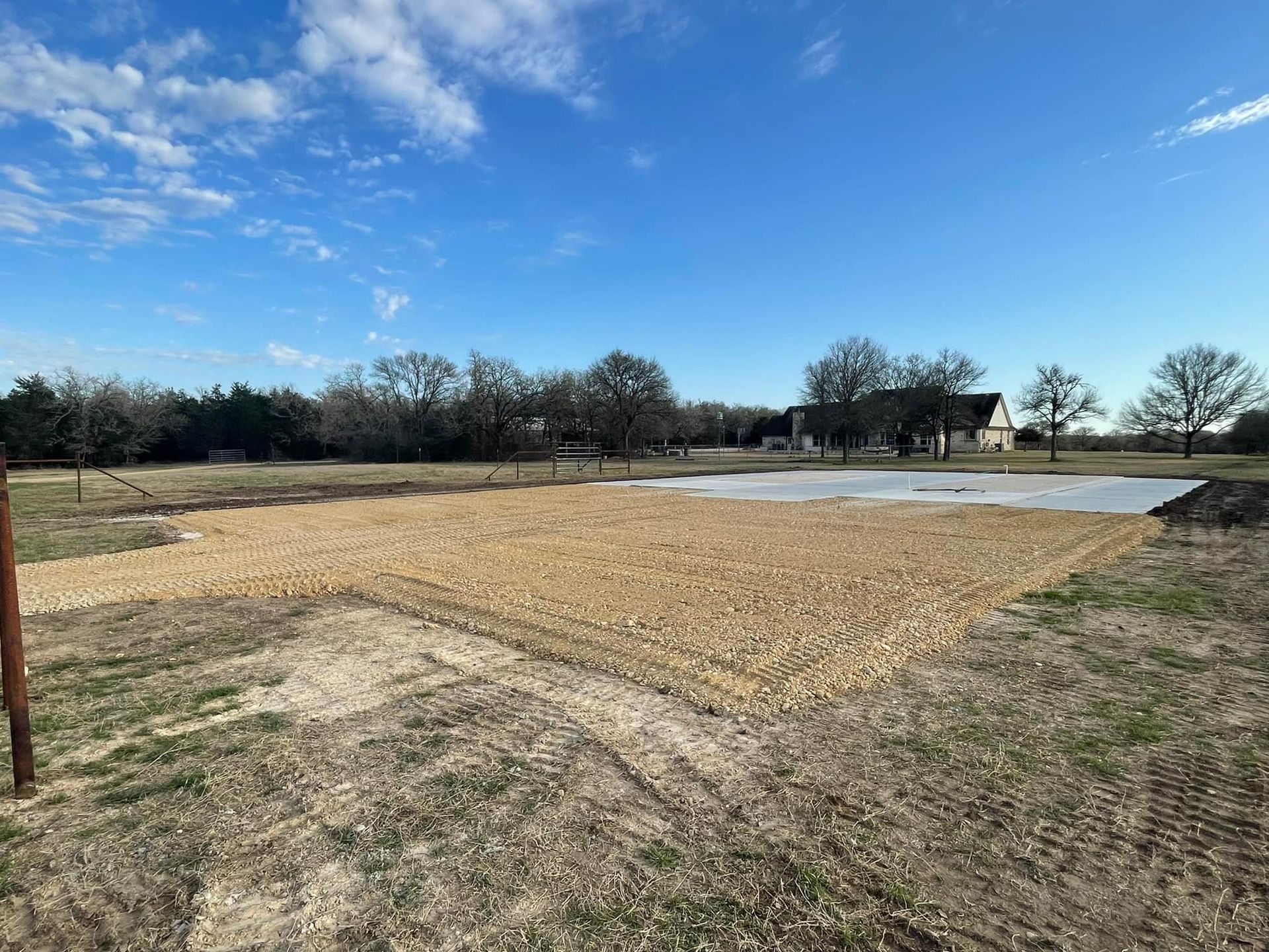 Gravel base for a structure on a grassy lot under a blue sky. Trees and a house are in the background.