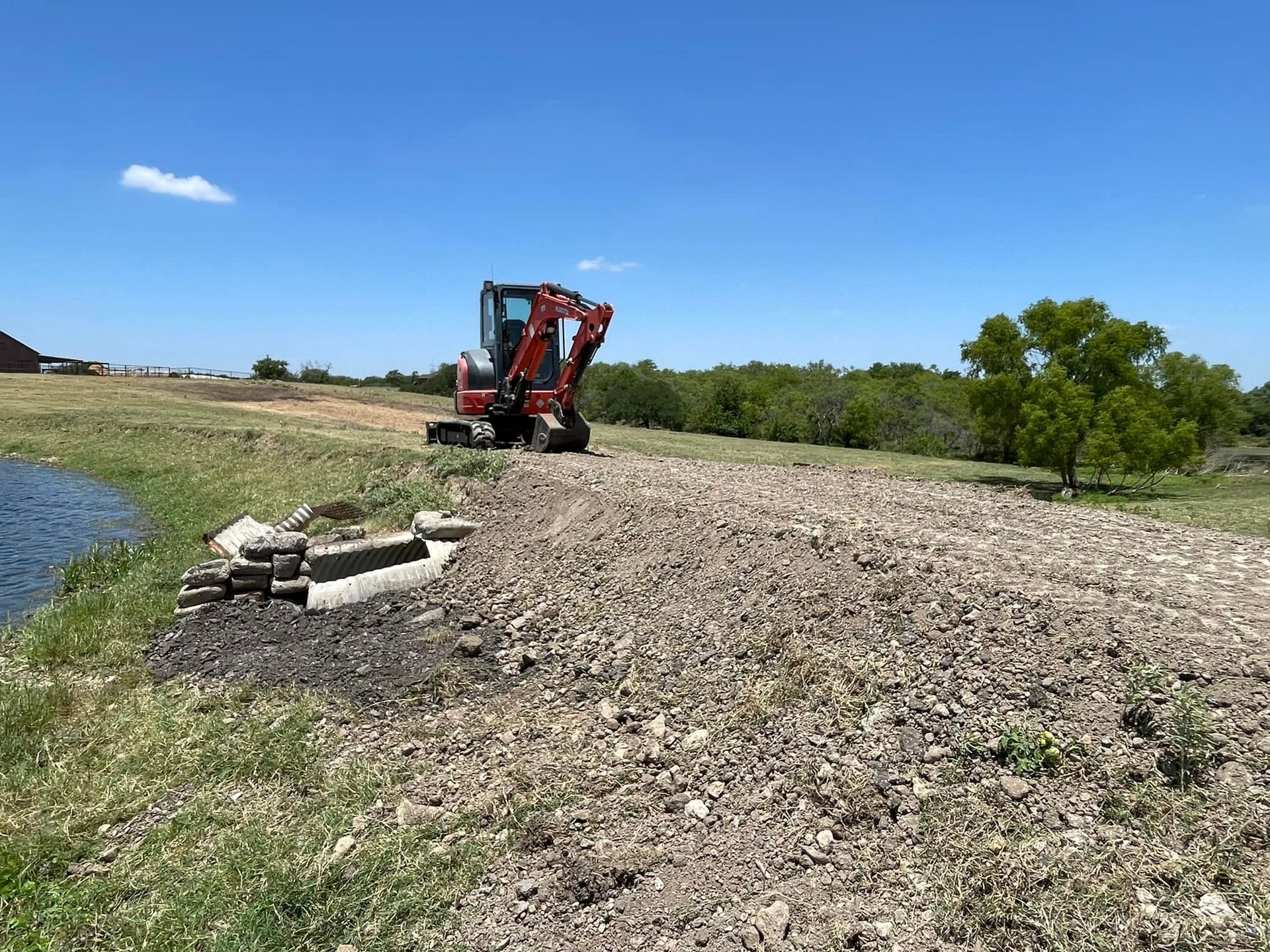 Red excavator on a gravel pile next to a pond, under a bright blue sky.