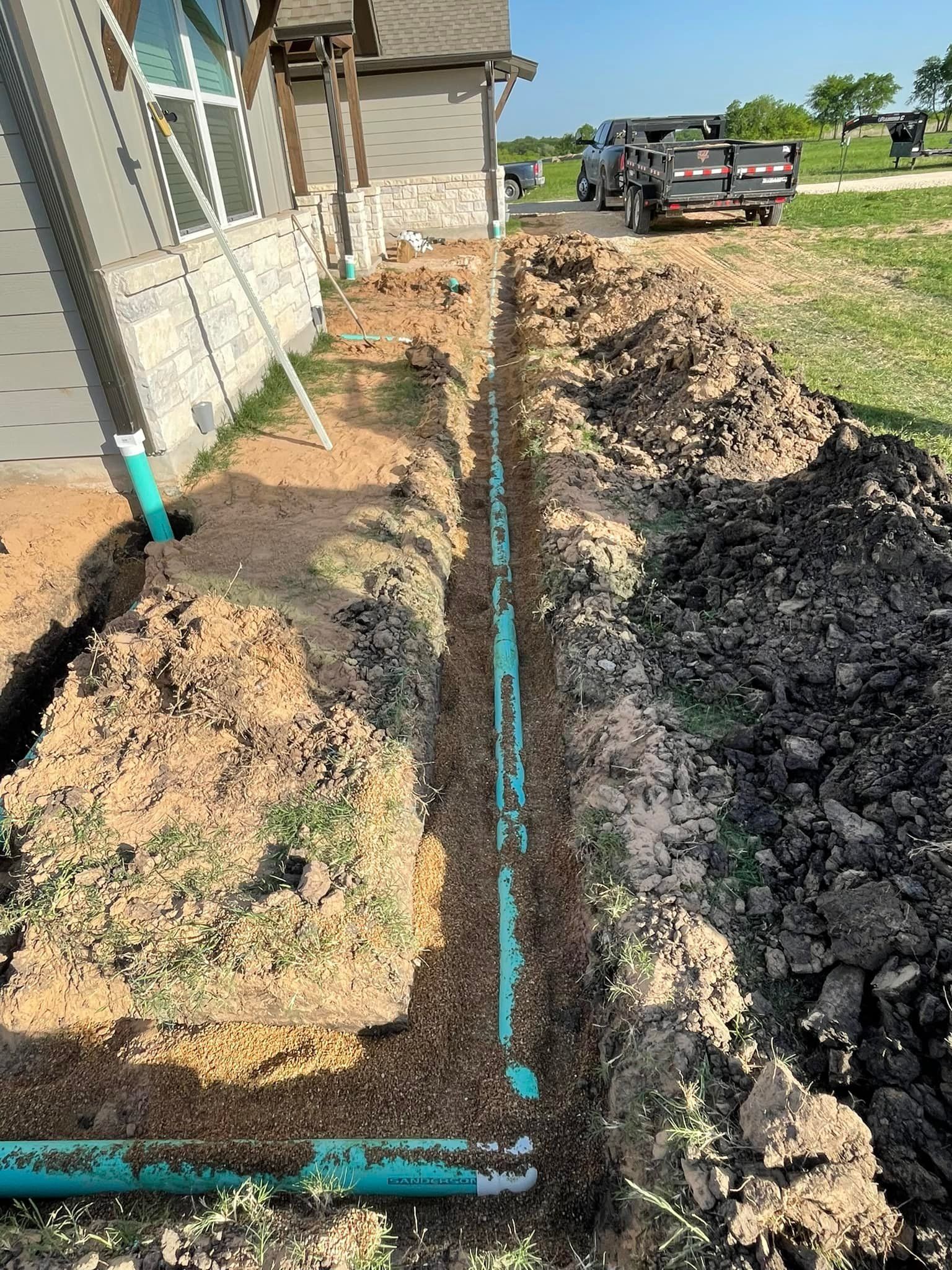 A trench dug alongside a house, revealing green pipes. Construction site.