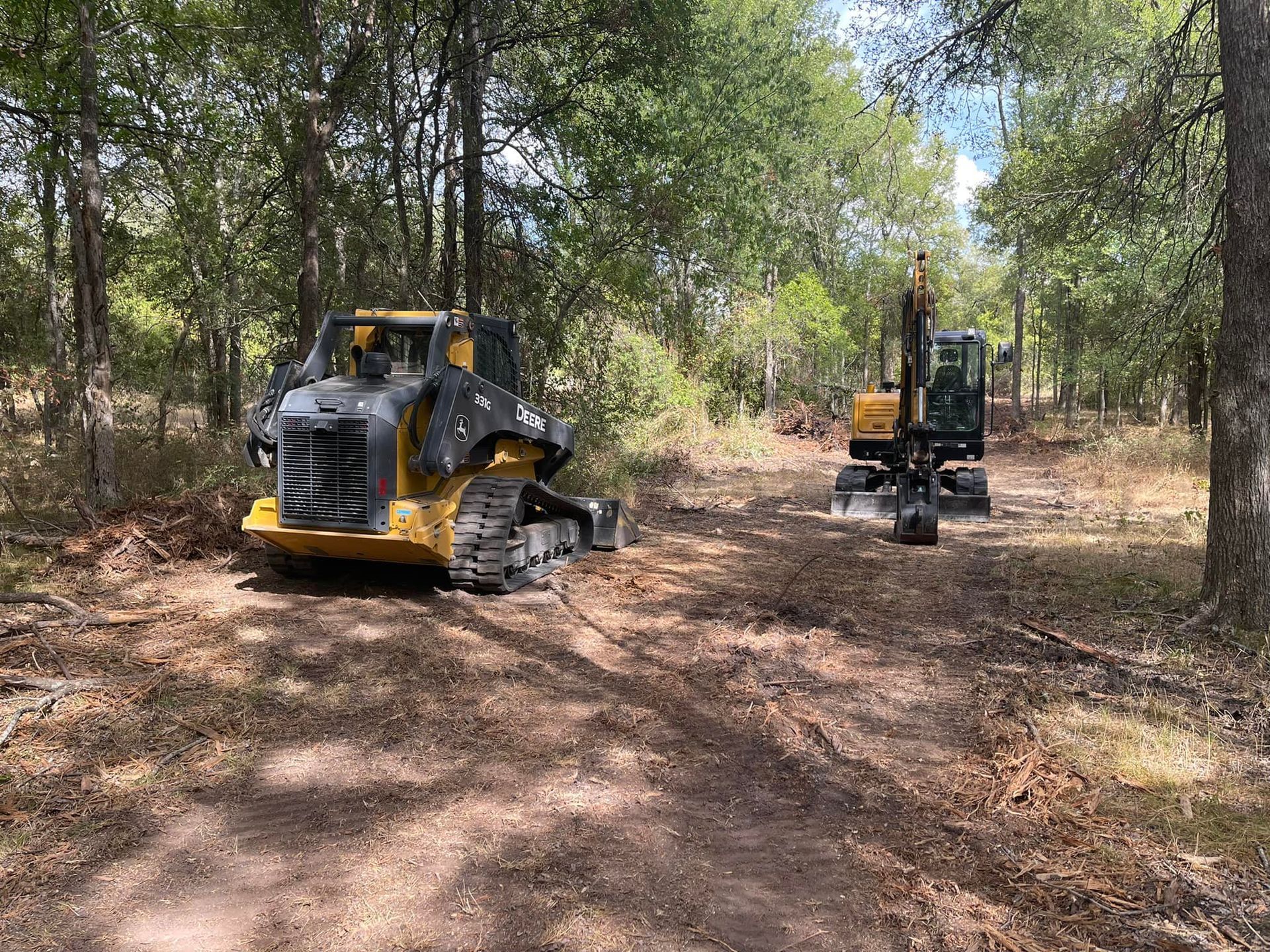 Two yellow construction vehicles on a dirt path in a wooded area.