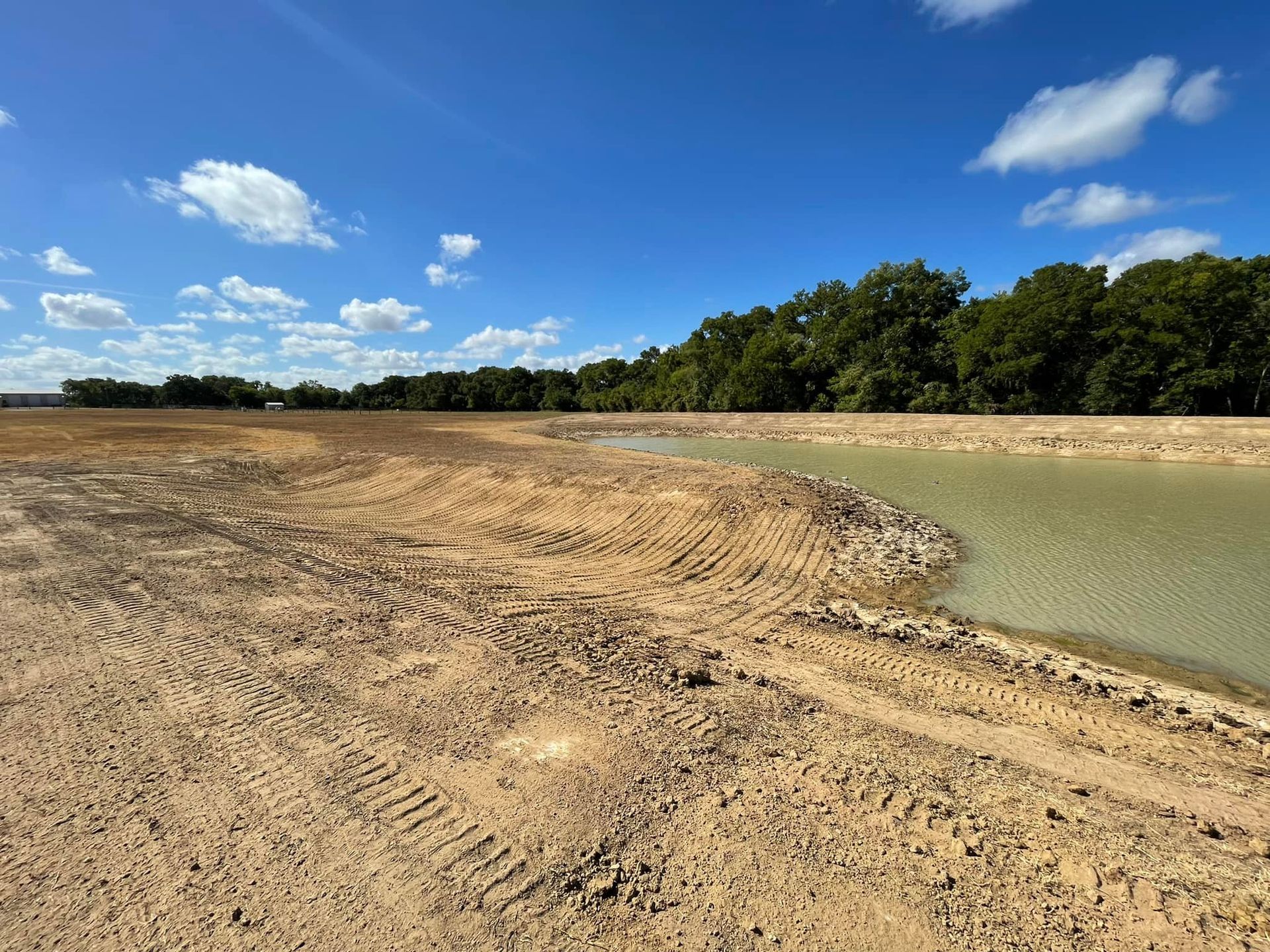 A barren, muddy construction site with a water-filled area and trees under a blue sky.