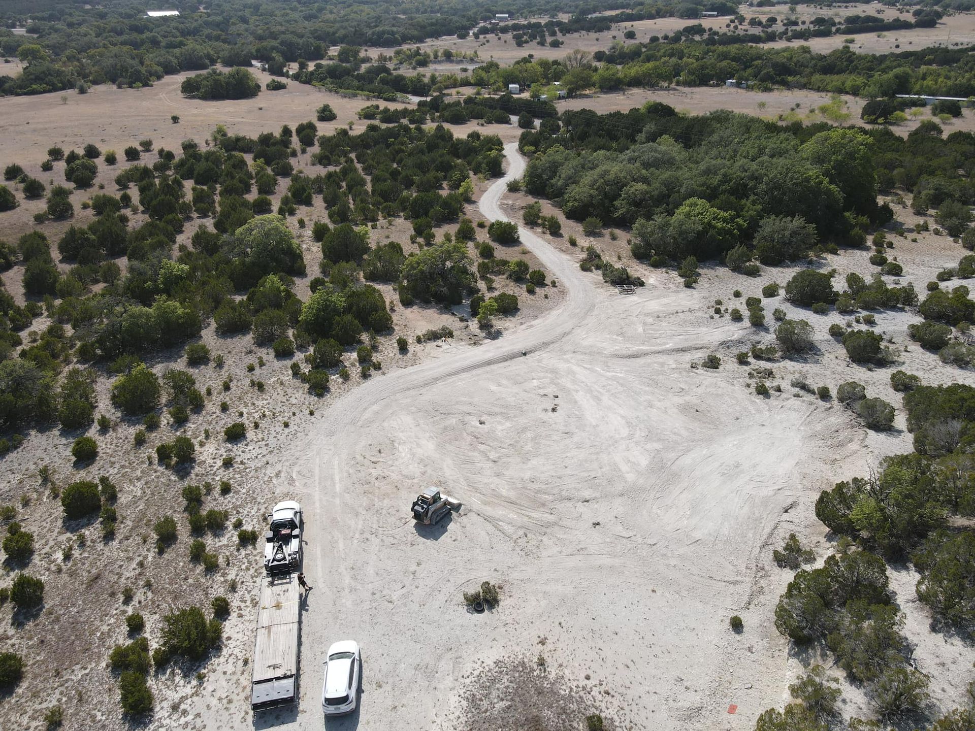 Aerial view of a dusty, cleared area with vehicles and a curving dirt road surrounded by trees and scrub.
