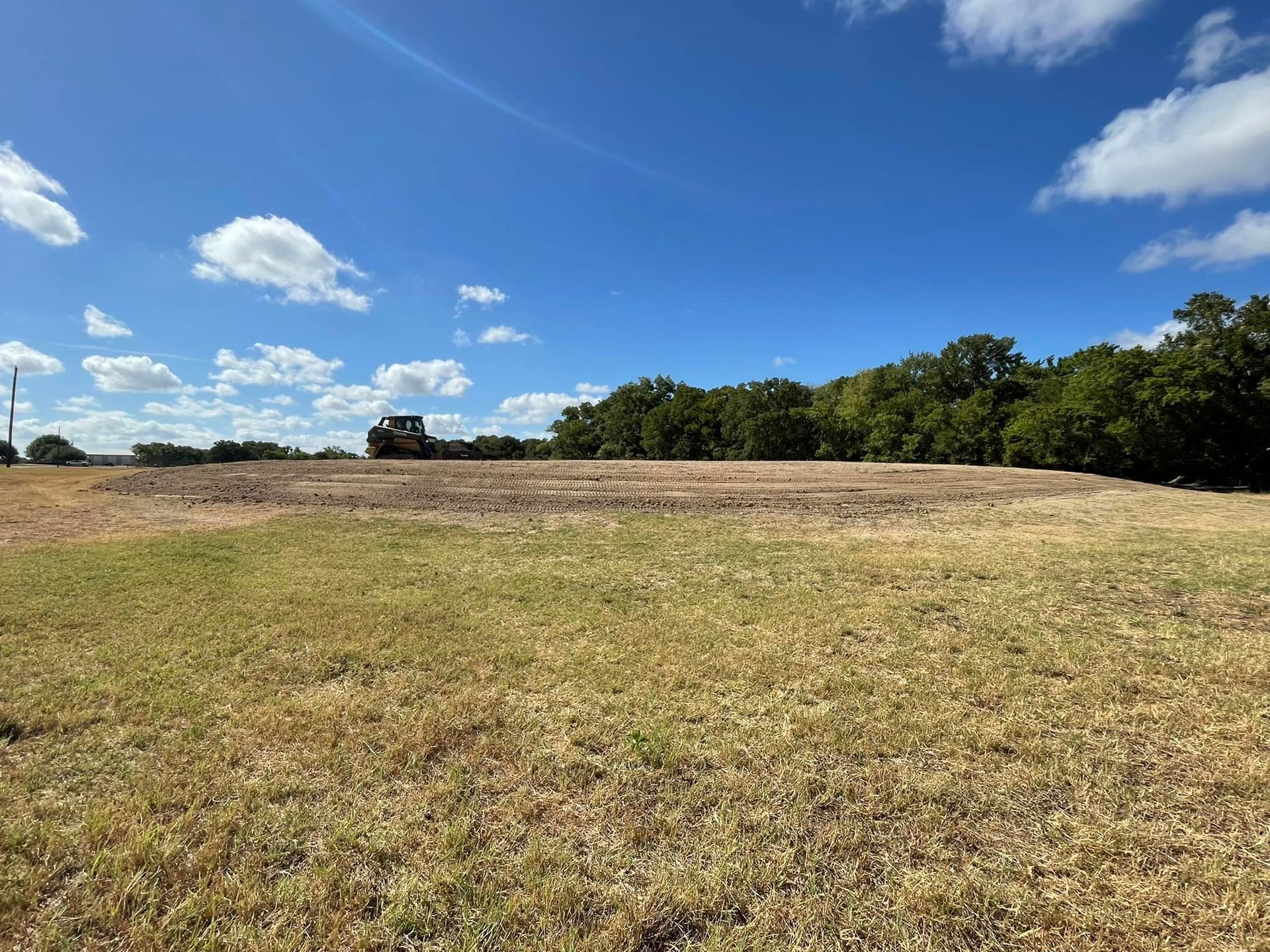 Grassy field with a cleared area, trees in the background, a bulldozer, and a blue sky with clouds.