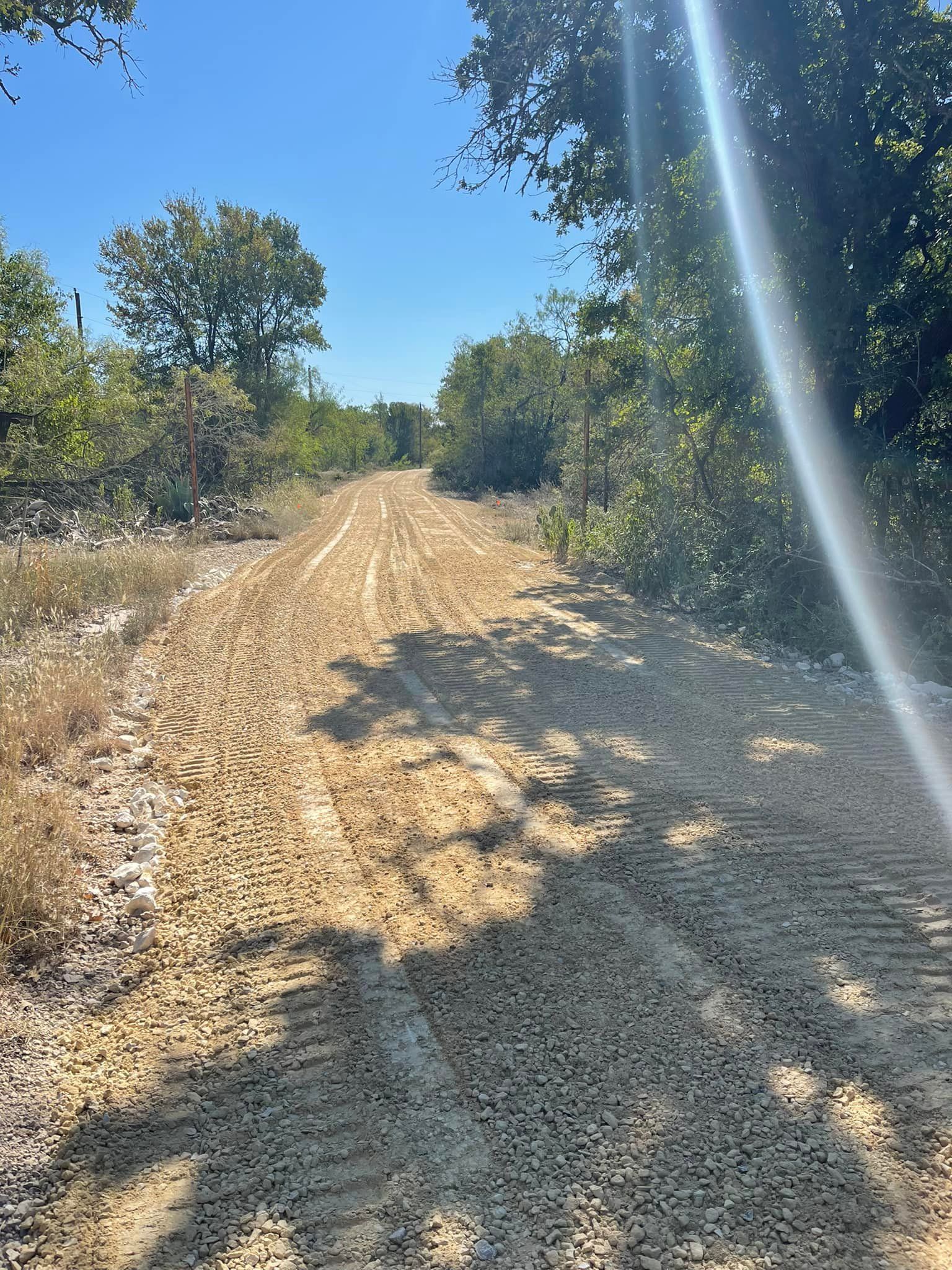 Gravel road through a wooded area on a sunny day. Tire tracks are visible on the road.