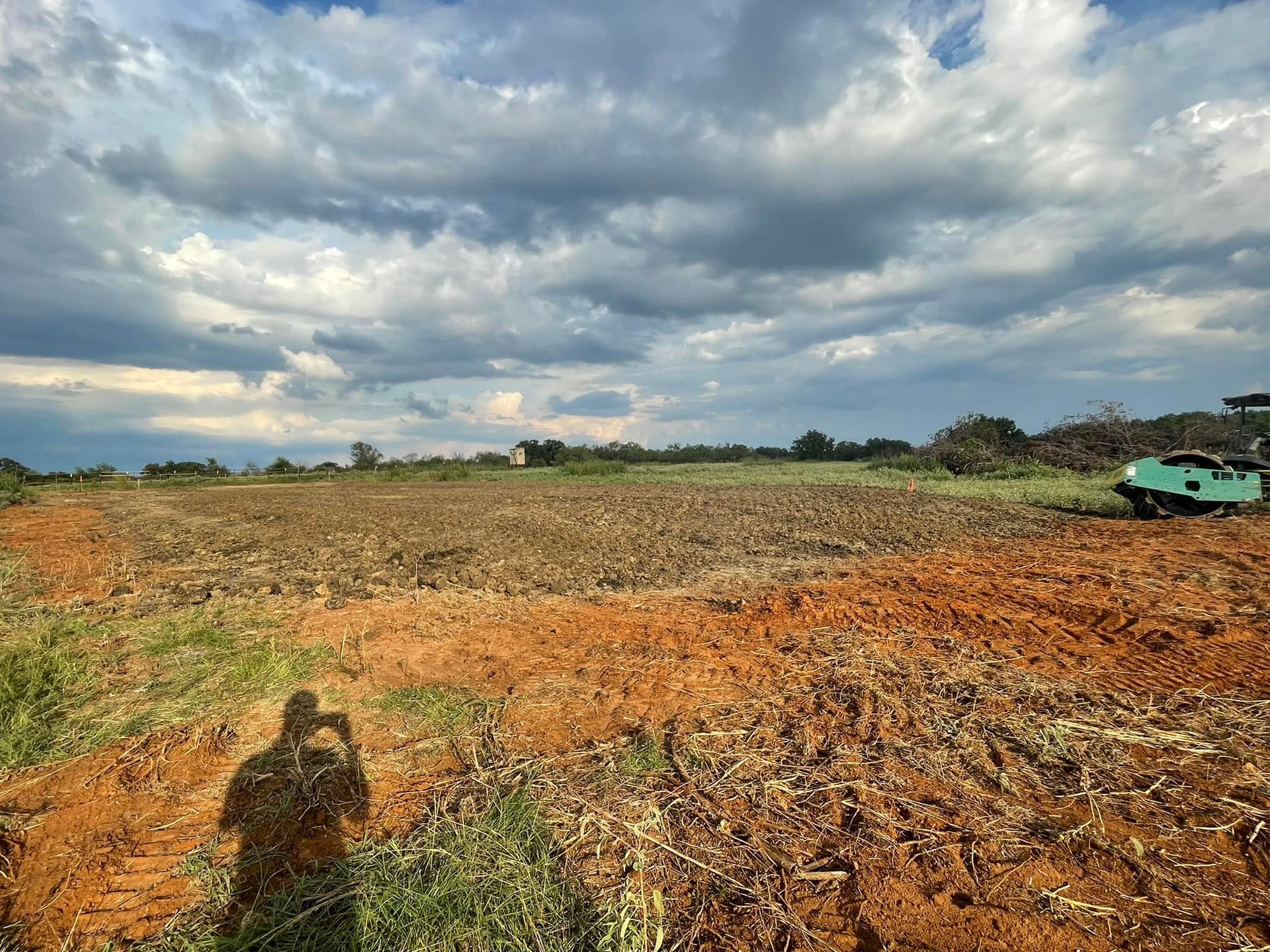 Tilled farmland under a cloudy sky; a tractor sits in the corner, a shadow of a person taking the photo.