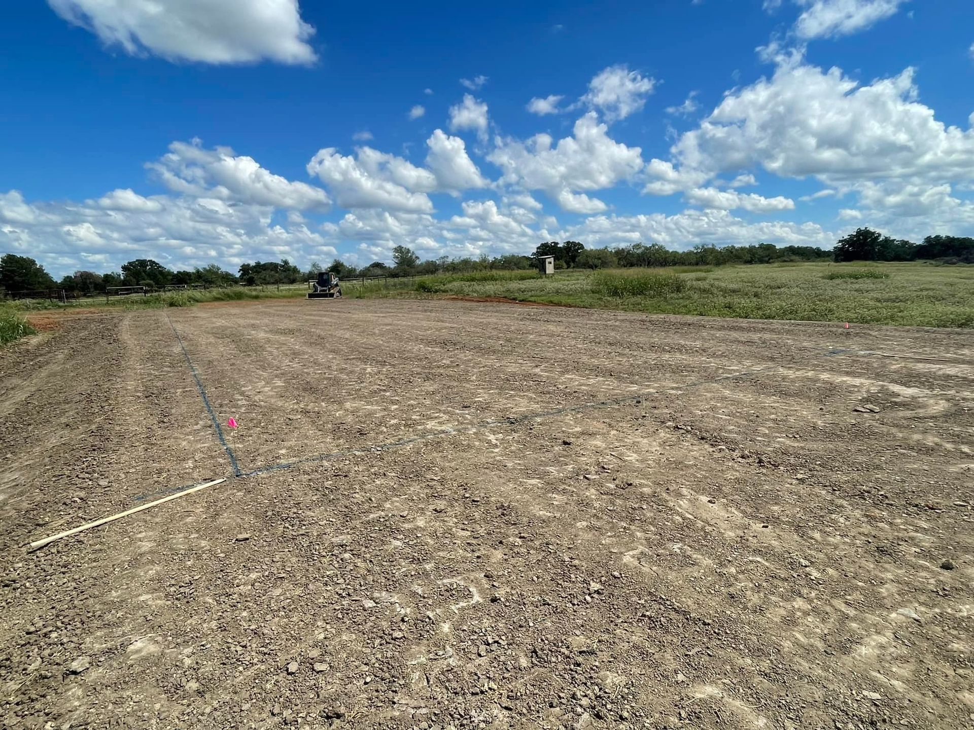 Dirt field under a bright blue sky with fluffy white clouds. Trees in the distance.