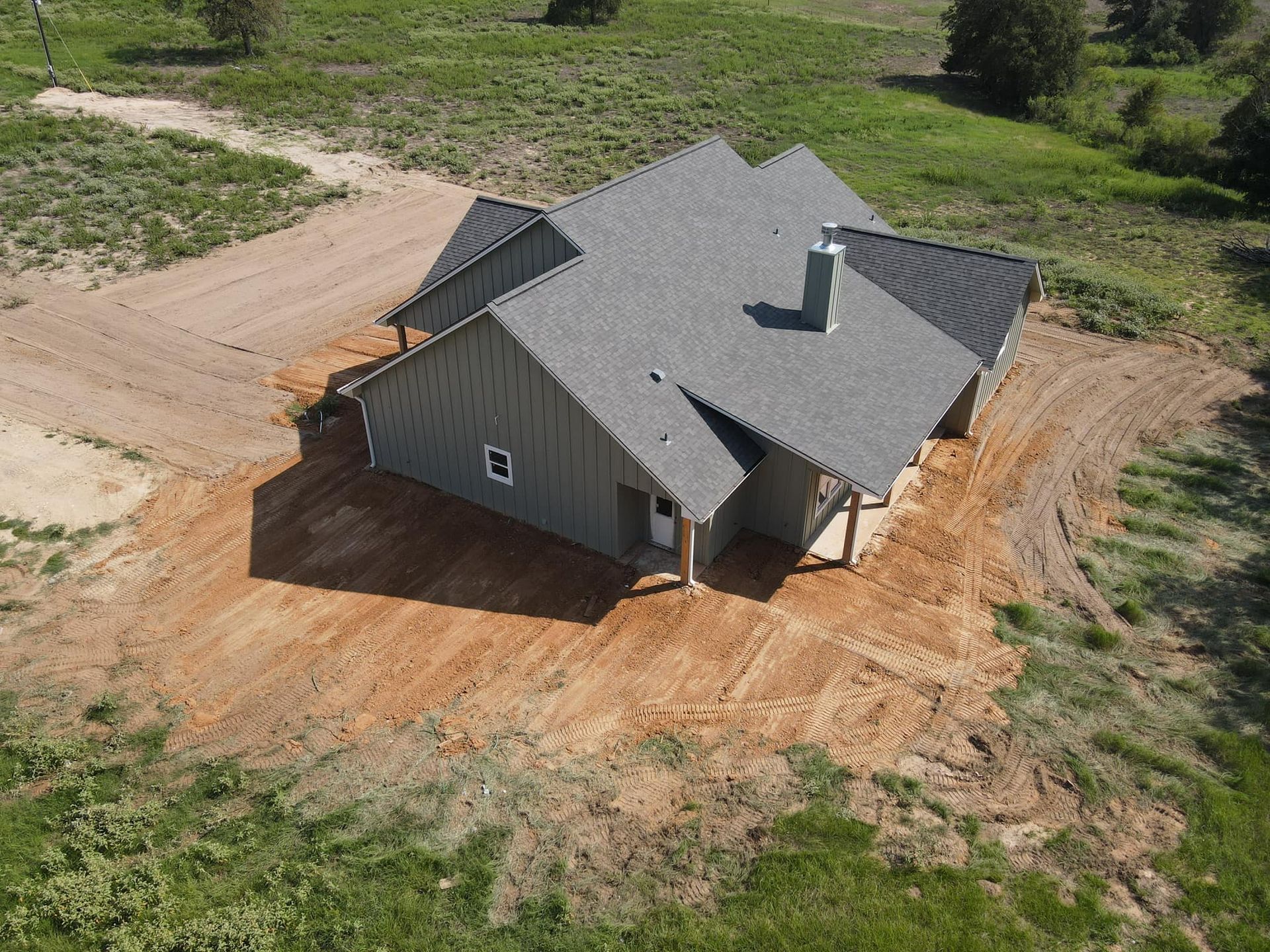 Gray house with dark roof and chimney on dirt patch surrounded by green grass.