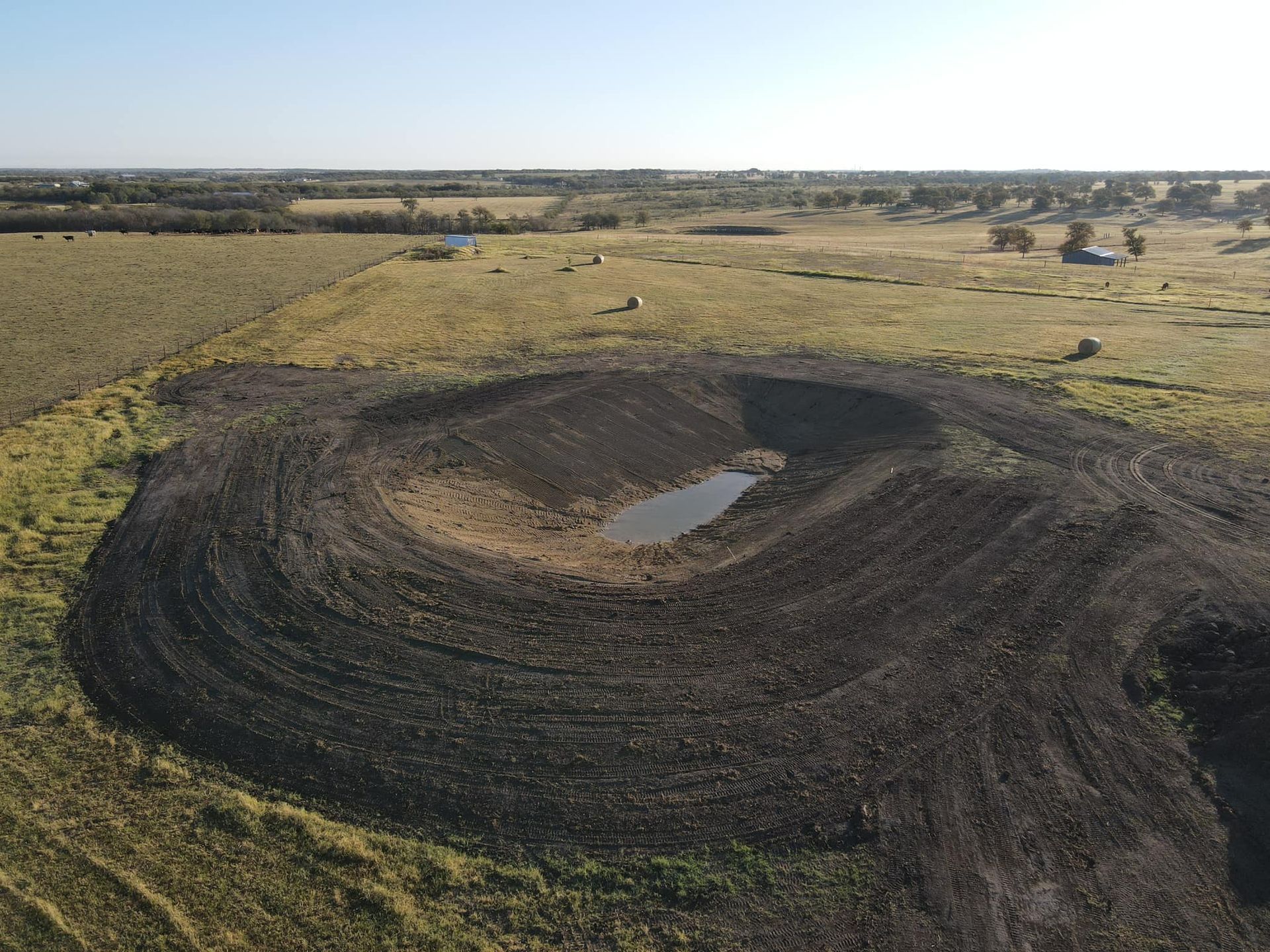 An aerial view of an excavated, circular pond with a small amount of water, surrounded by open fields.