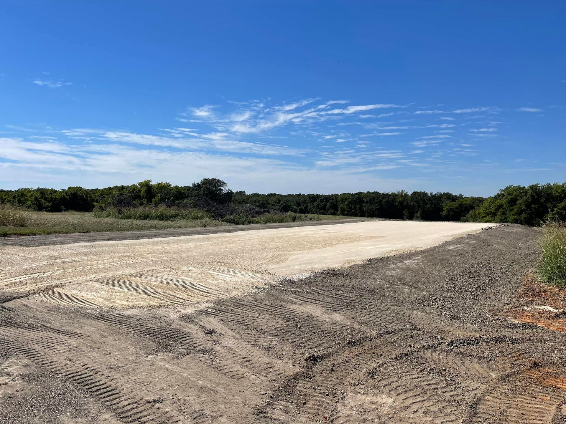 Open field with light-colored sediment in the center, surrounded by darker soil. Trees line the horizon, under a blue sky.