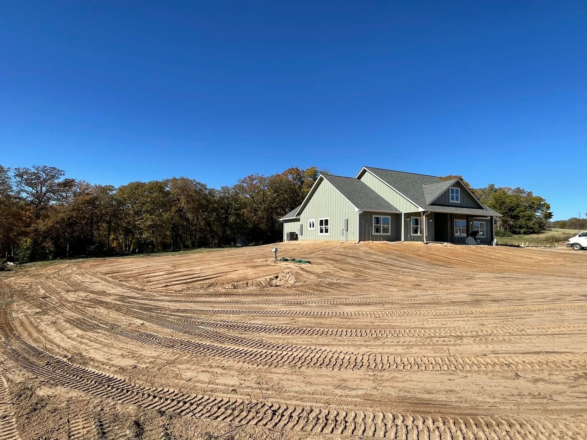 A new house under construction sits on a leveled, sandy lot with tire tracks, under a clear blue sky.