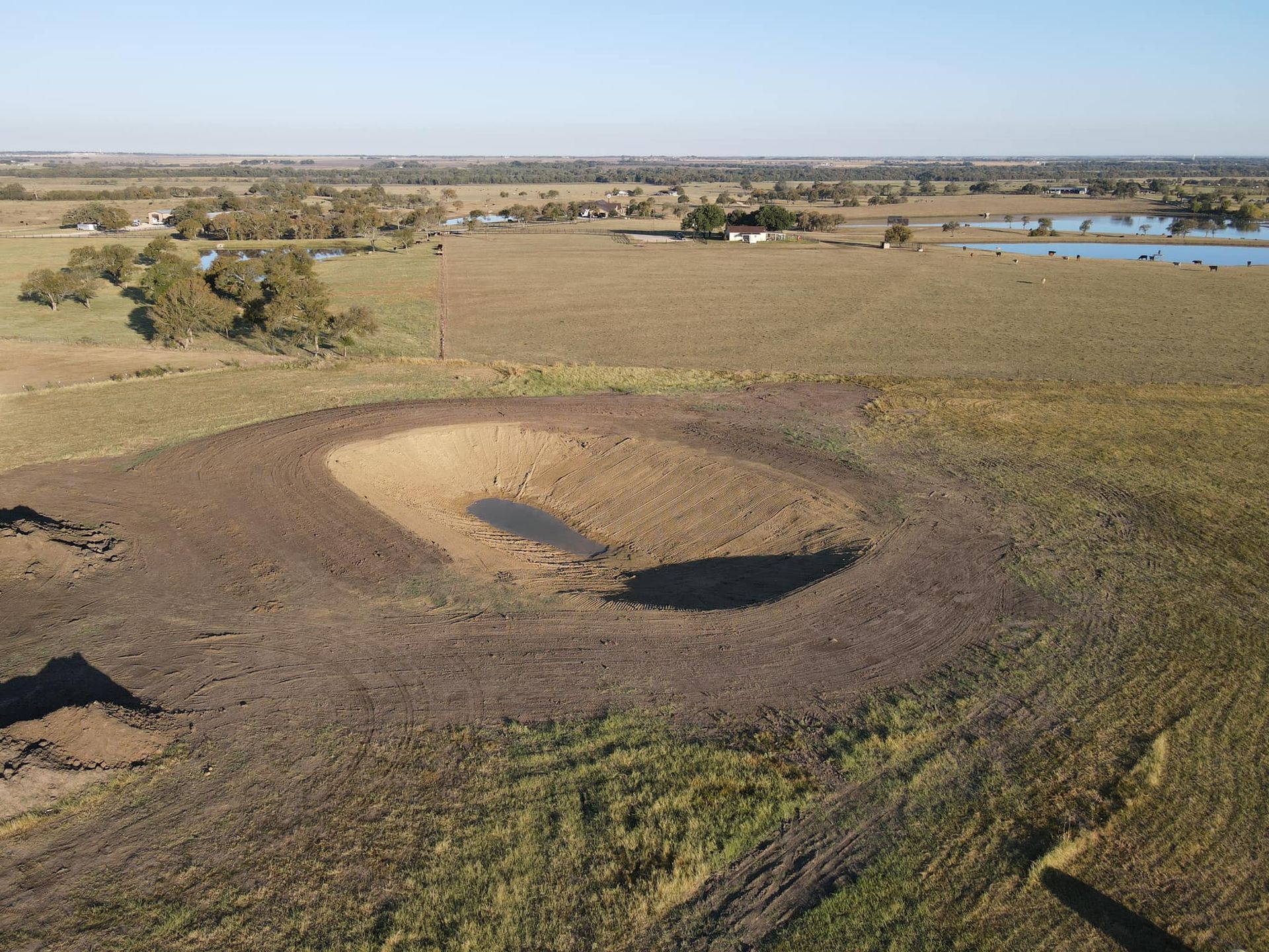 Aerial view of a large, dry, circular watering hole in a brown grassy field under a blue sky.