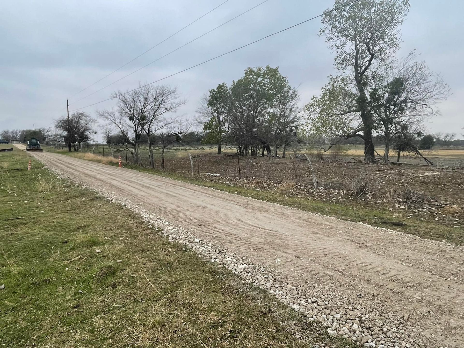 Gravel road through rural landscape with trees and power lines under an overcast sky.