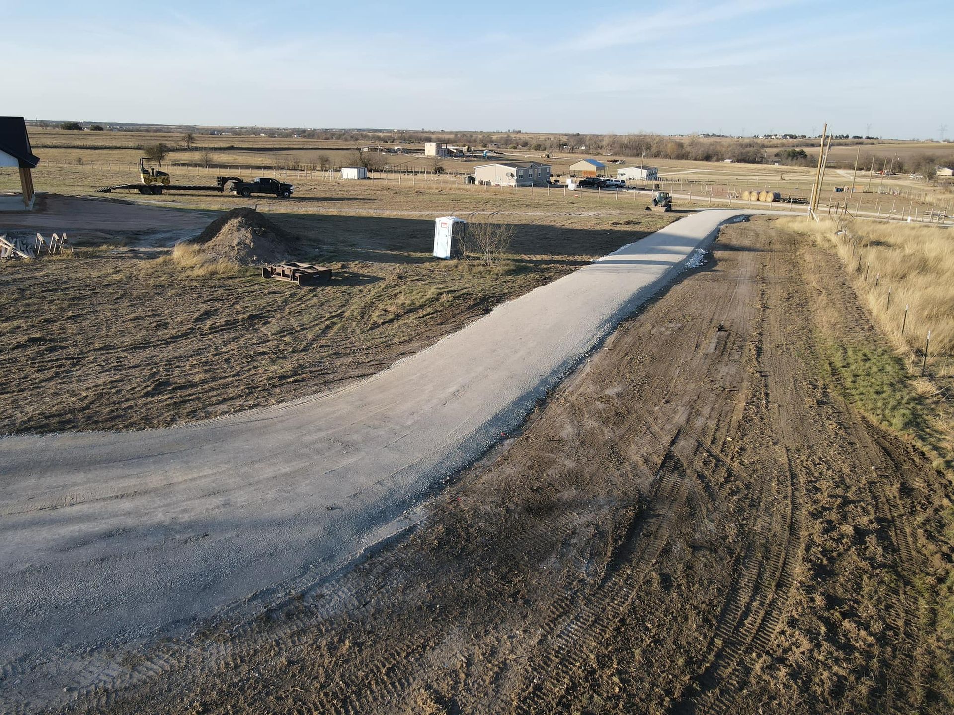 A gravel road leads through a brown field, with construction equipment and structures in the background under a blue sky.