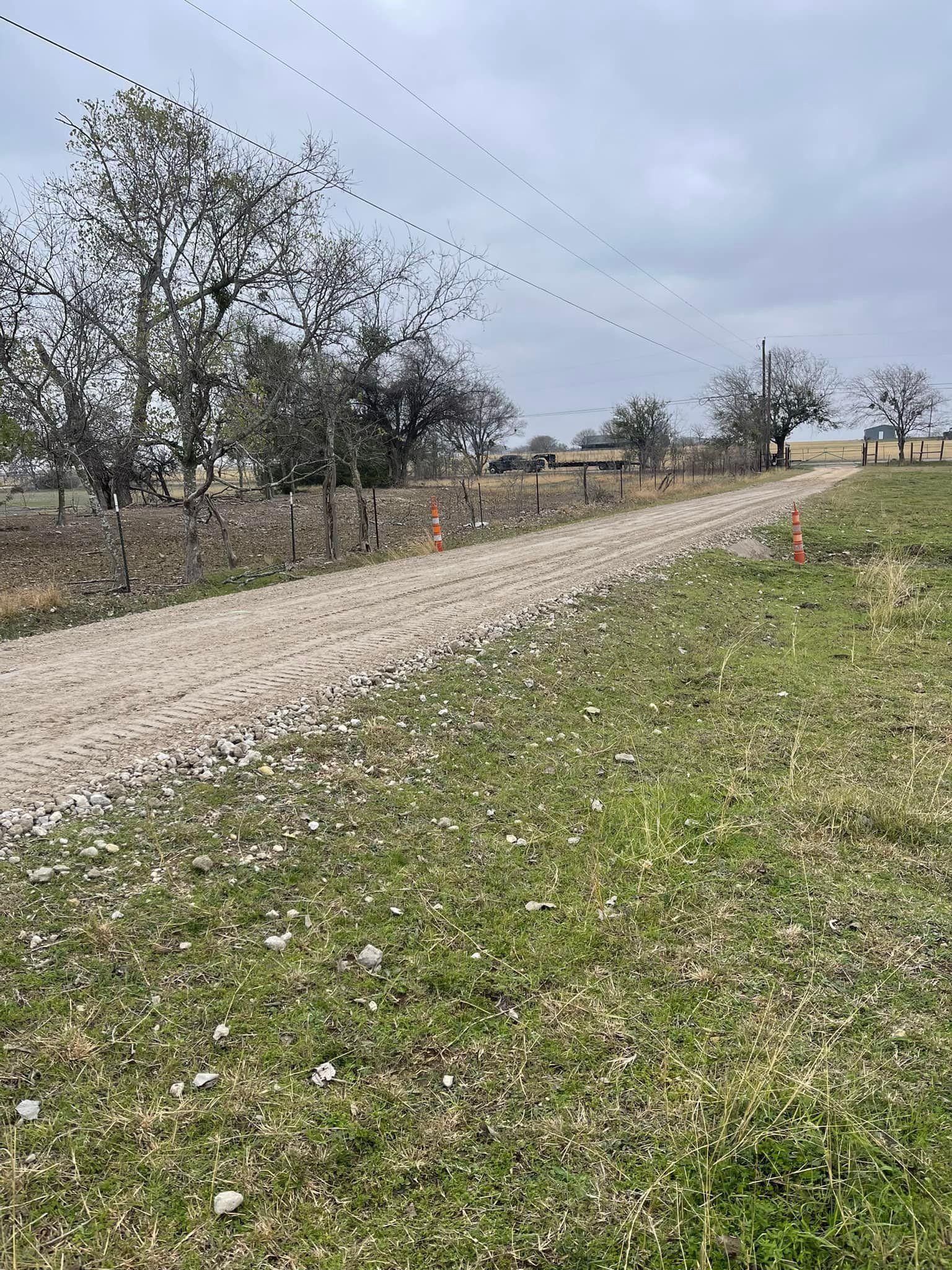 Gravel road through grassy field, fence and trees along the side, overcast sky overhead.