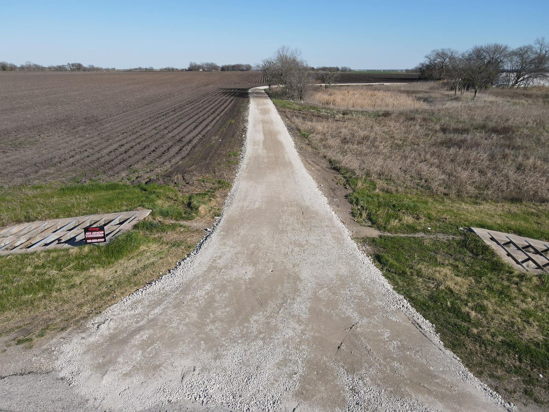 Gravel path through fields under blue sky, with grassy edges and bare trees in the distance.