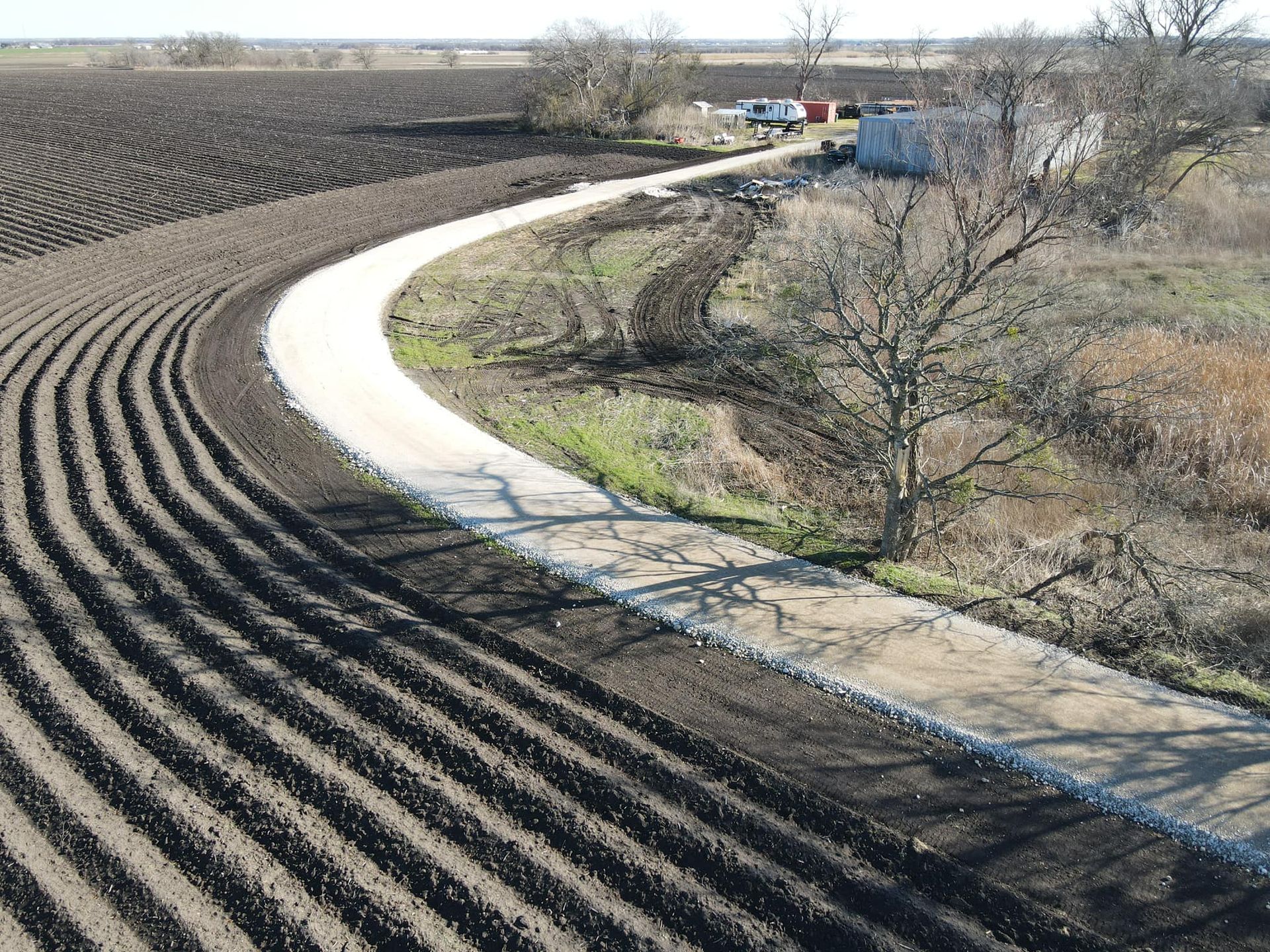 Dirt road curves through plowed field, with building and trees in the background.