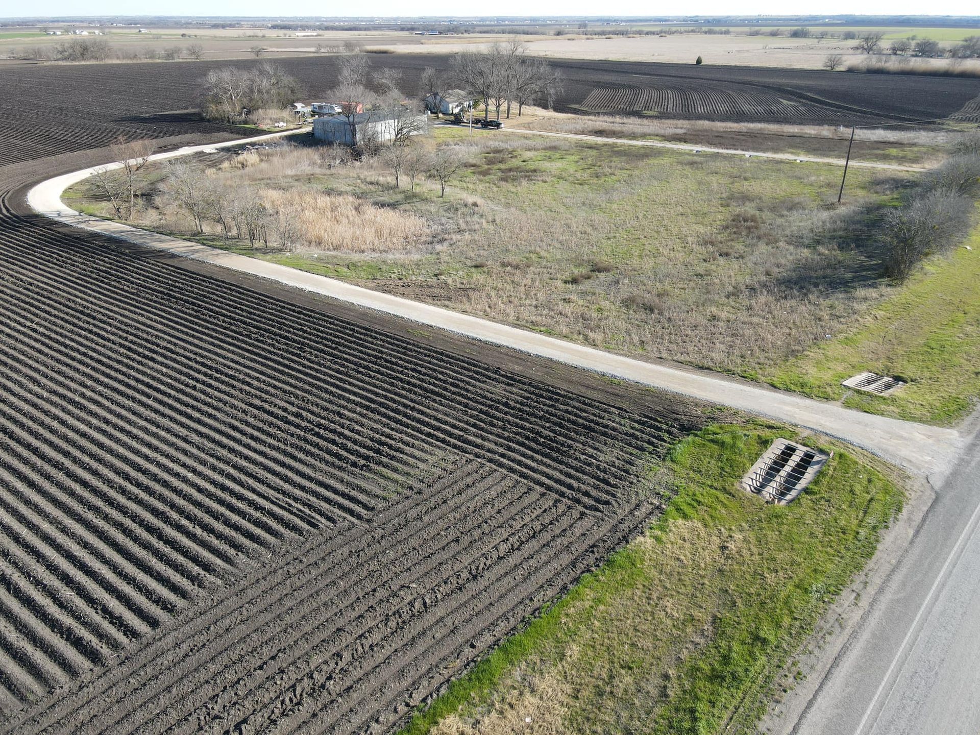 Fields of dark earth with a long road leading to a house and a grassy patch.