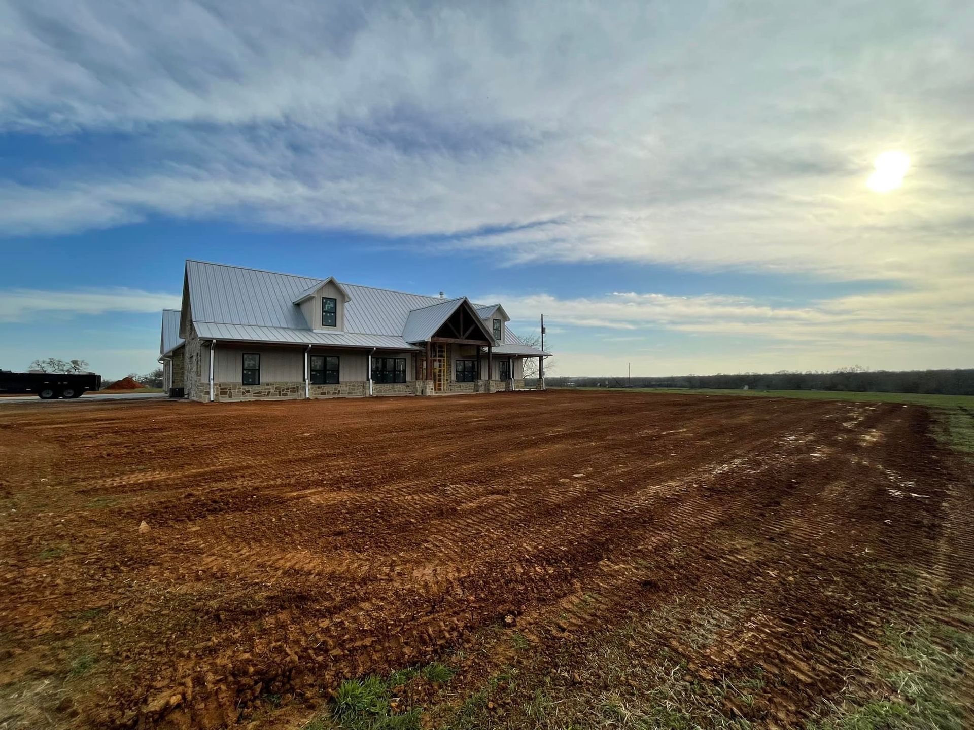 A large, two-story home with a stone exterior sits on a recently tilled field under a cloudy sky.