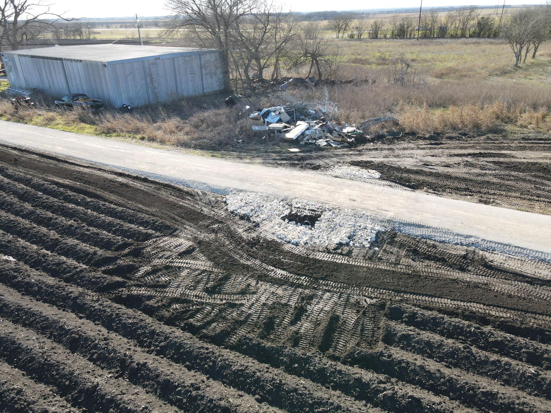 Dirt road damaged, crosses cultivated field, debris pile next to a shed in a rural landscape.