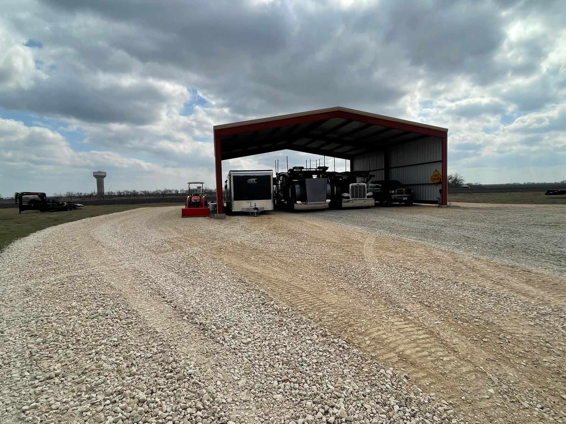 A gravel driveway leading to an open-sided metal shed. Construction equipment is parked inside and nearby. Cloudy sky.