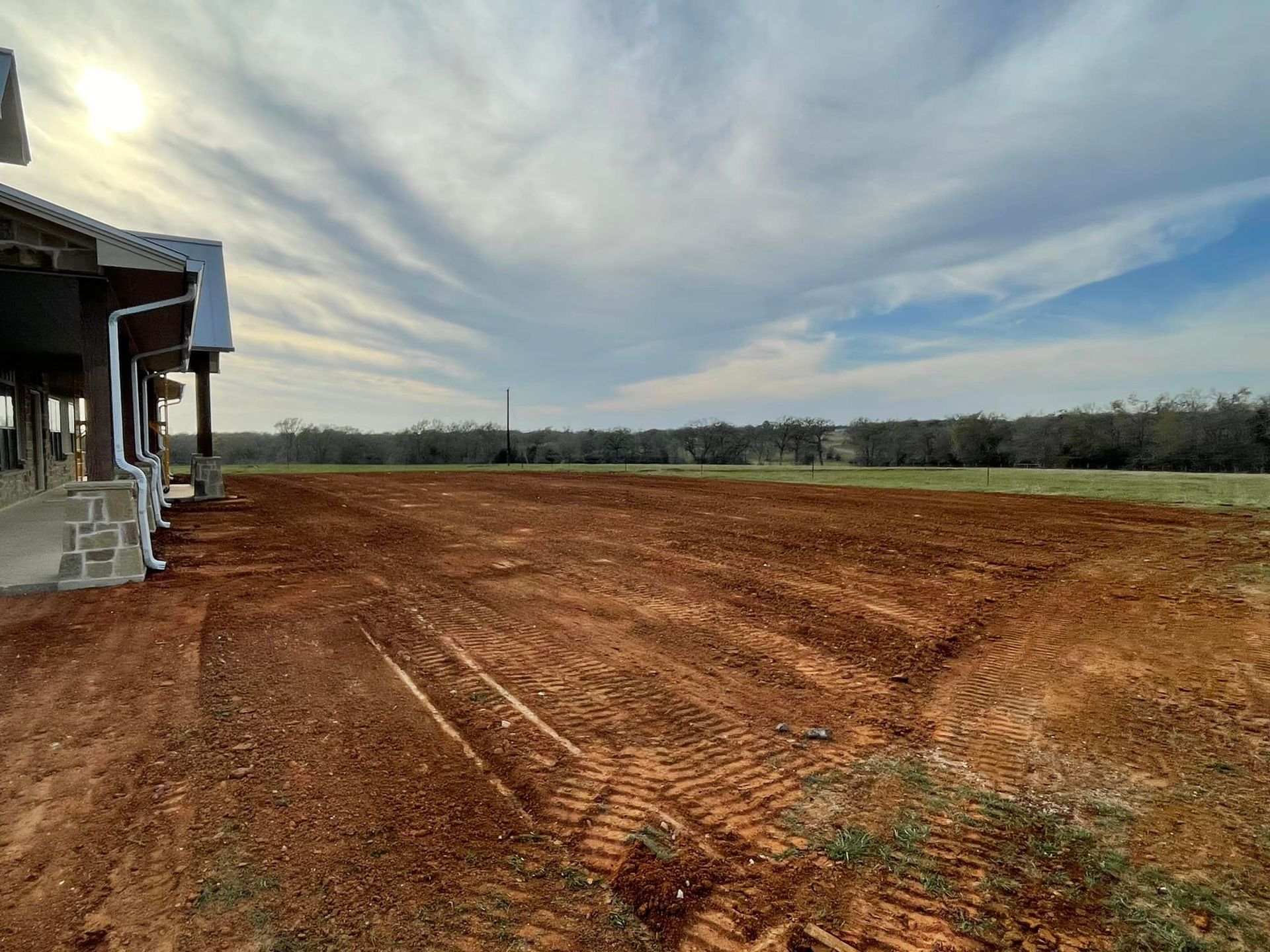 A freshly graded red dirt lot next to a building, under a cloudy blue sky.