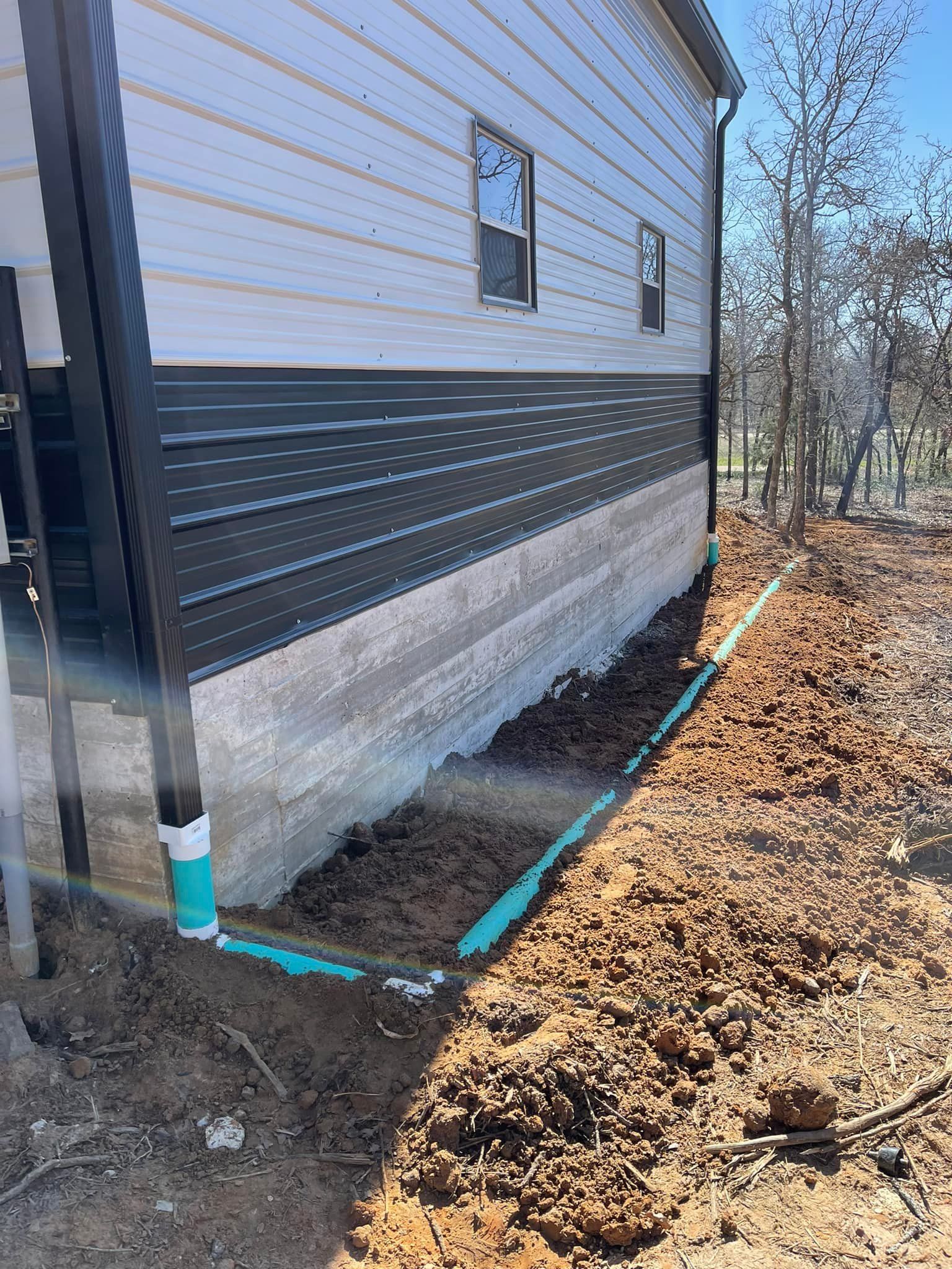 Blue pipes run along the foundation of a house under construction in a dirt lot.