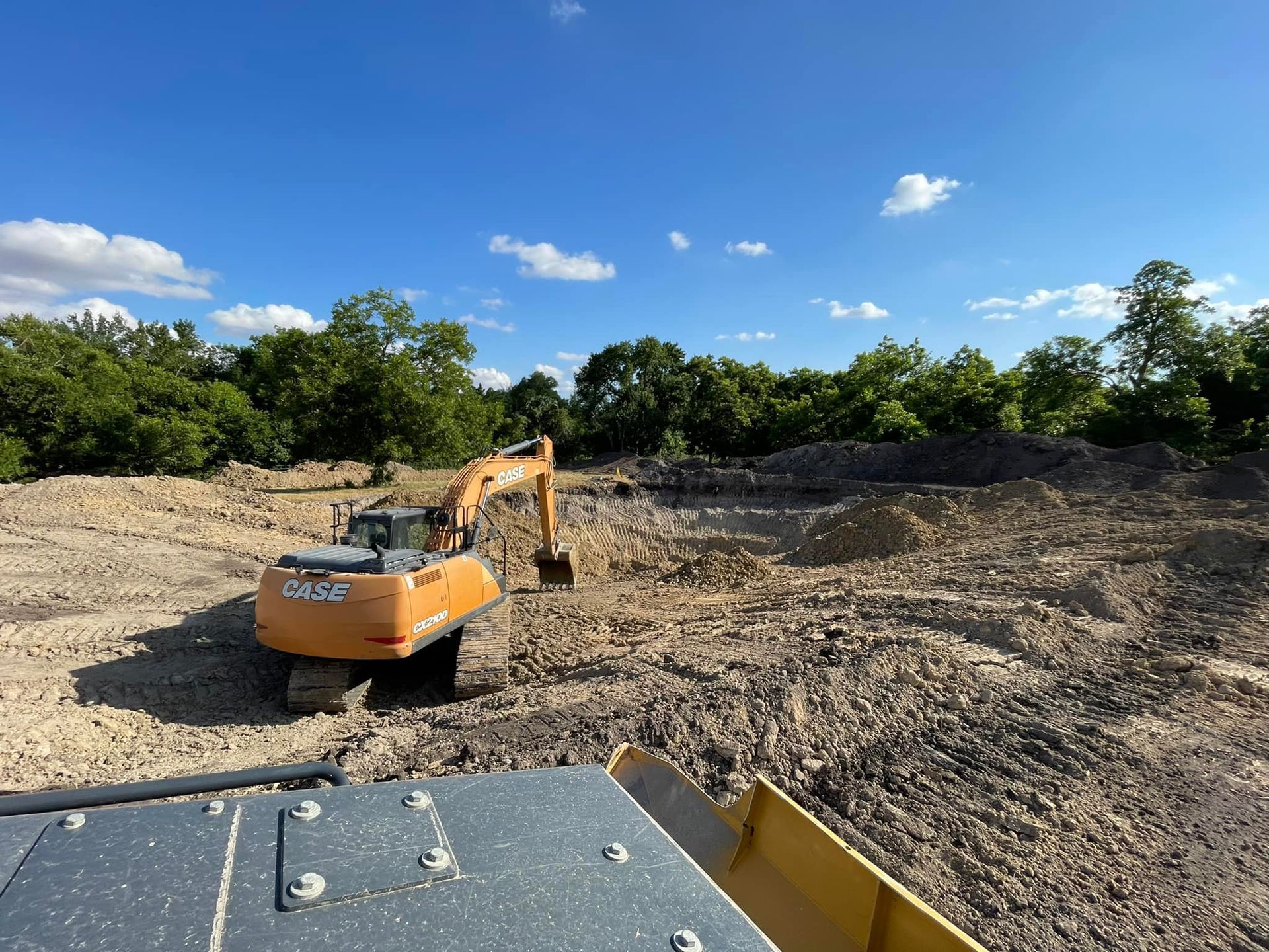 An orange excavator digs in a dirt field under a blue sky with trees in the background.