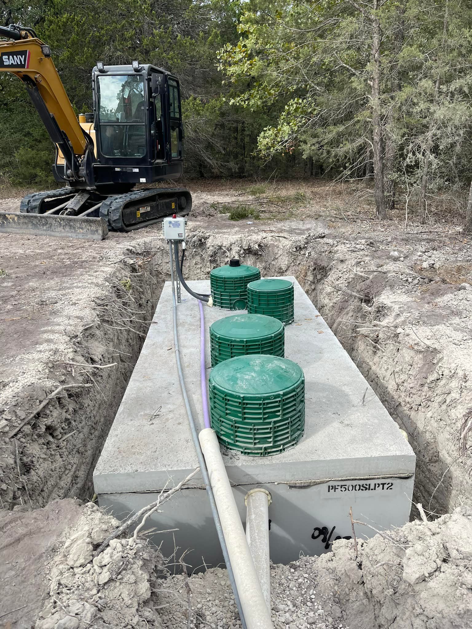 Excavated concrete septic tank with green access lids, next to a small excavator in a wooded area.