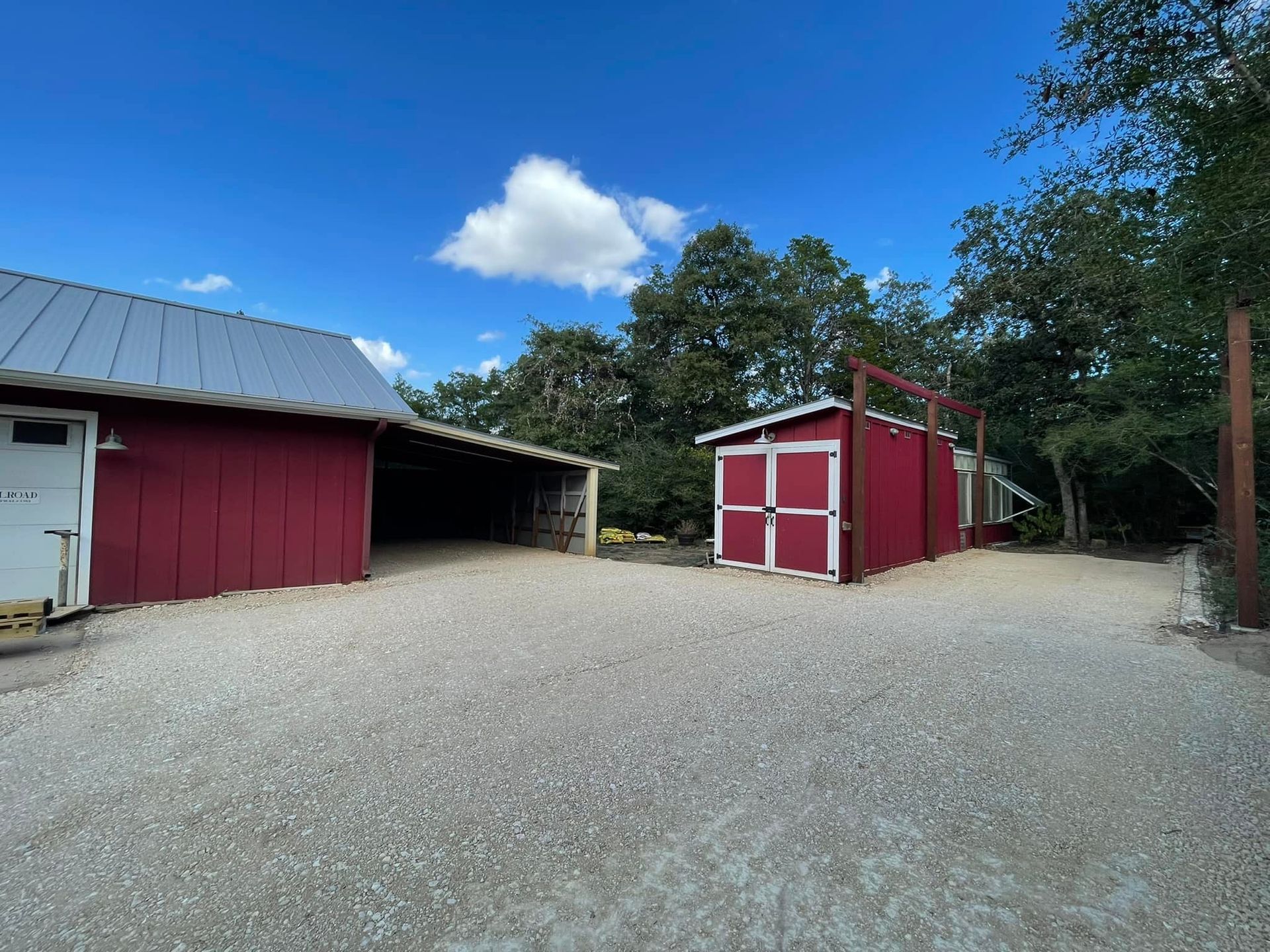 Red barn buildings on a gravel driveway under a blue sky with trees.