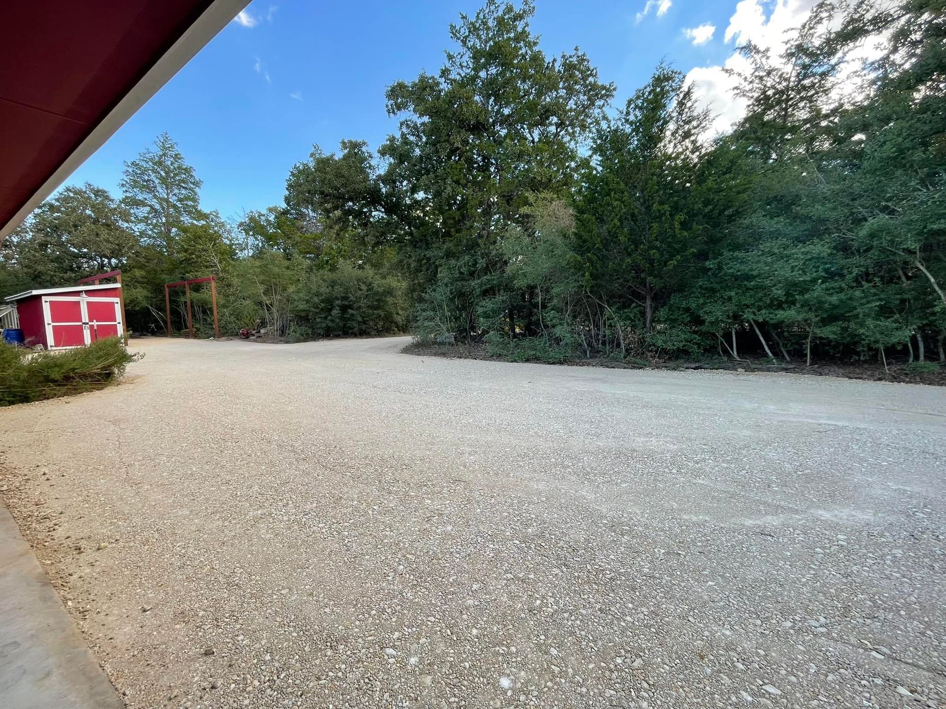 Gravel driveway leads to a red shed and lush trees under a blue sky.