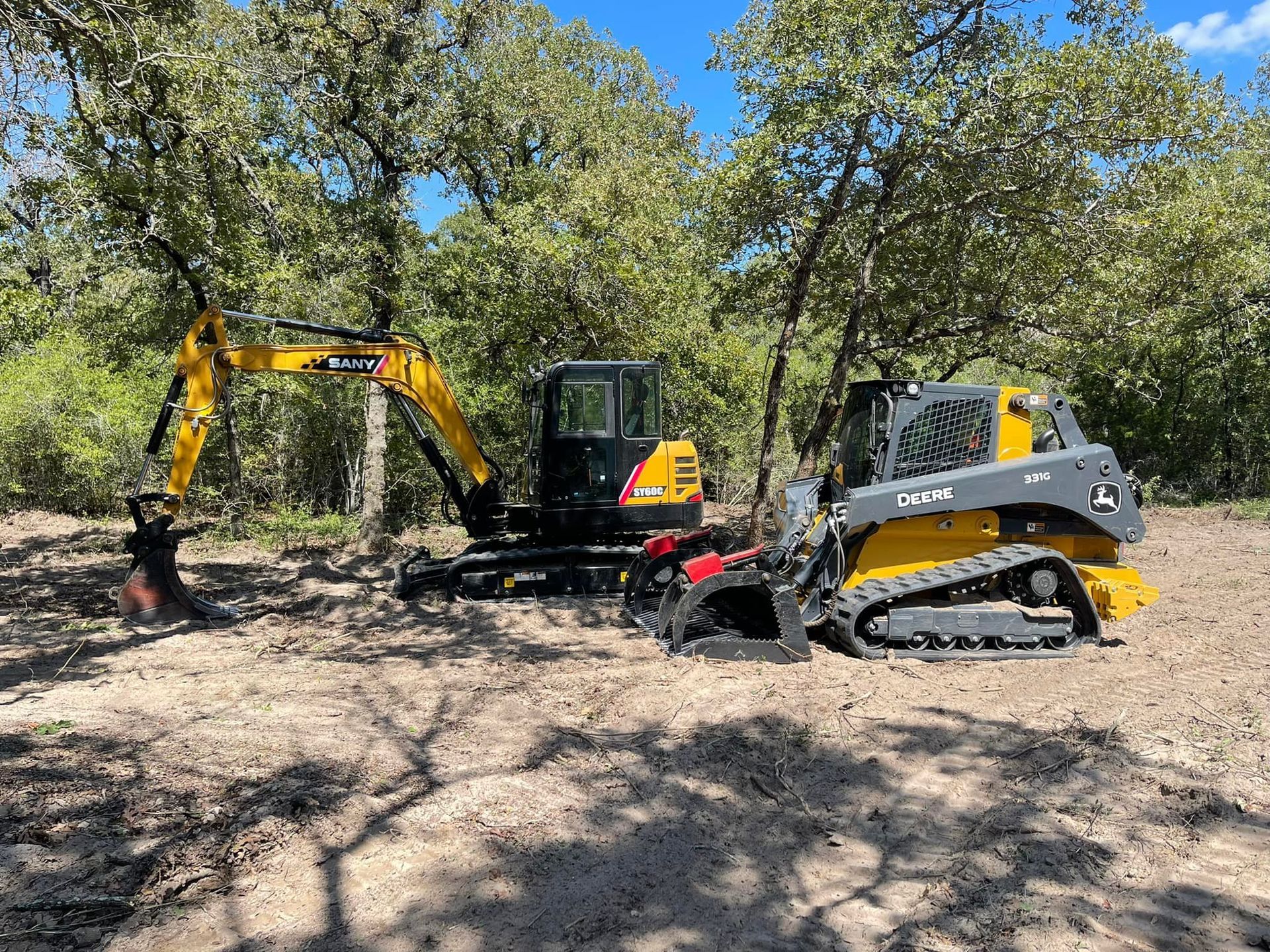 Yellow excavator and track skid steer on dirt clearing, trees in the background.