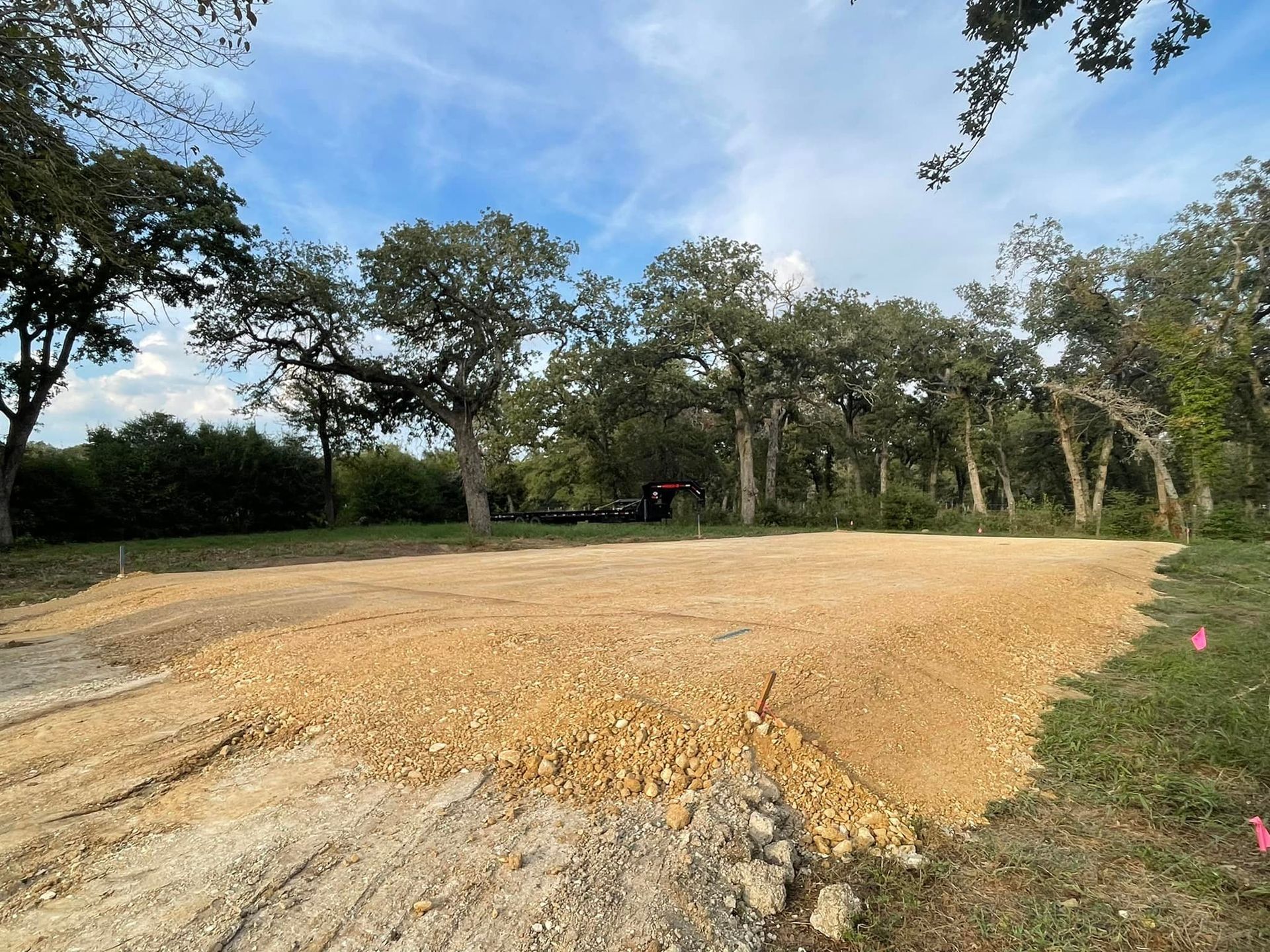 Clearing for construction. Brown gravel leveled in grassy area, trees in background, blue sky.