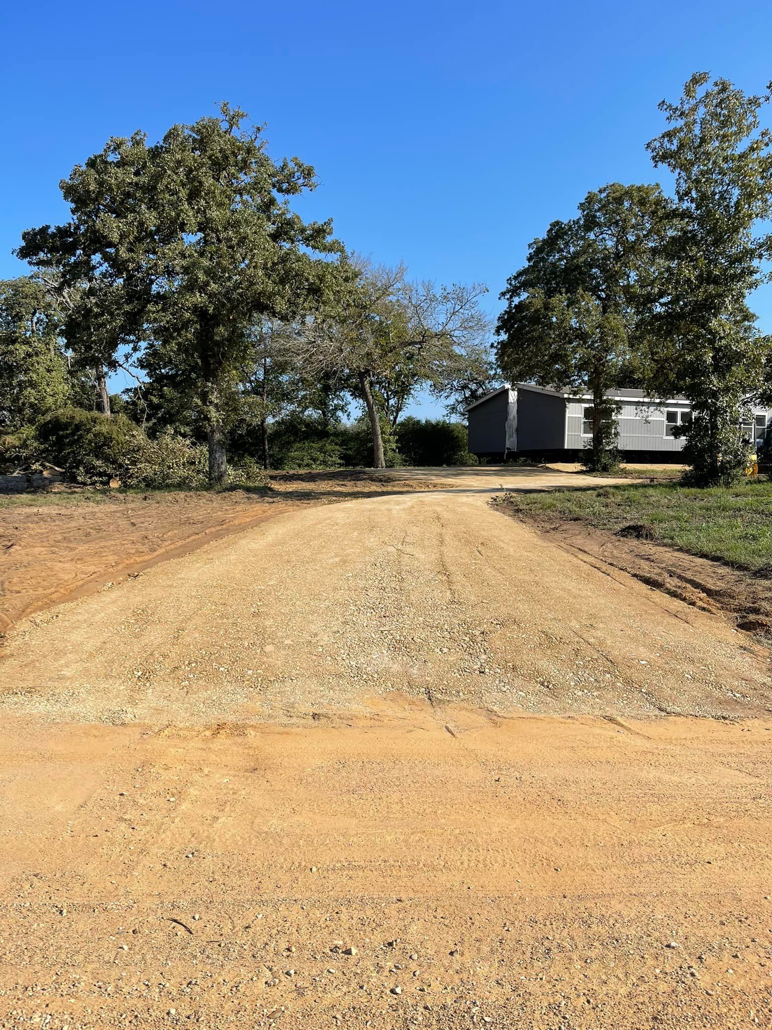 Gravel driveway leading to a trailer and trees under a bright blue sky.