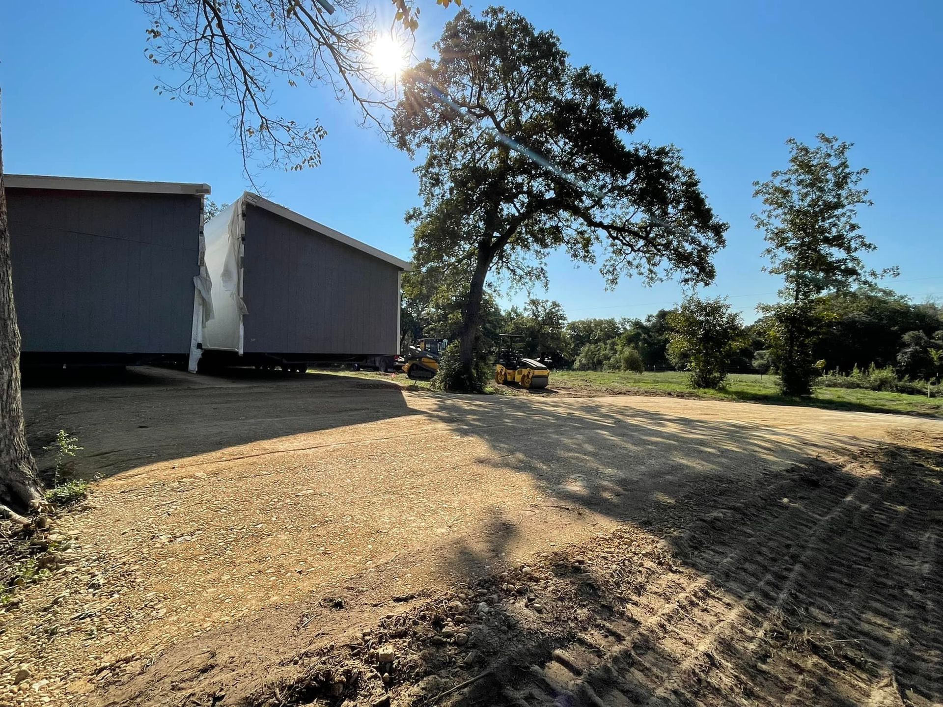 Two gray buildings on trailers in a field, a large tree, sunny sky, and construction vehicle visible.