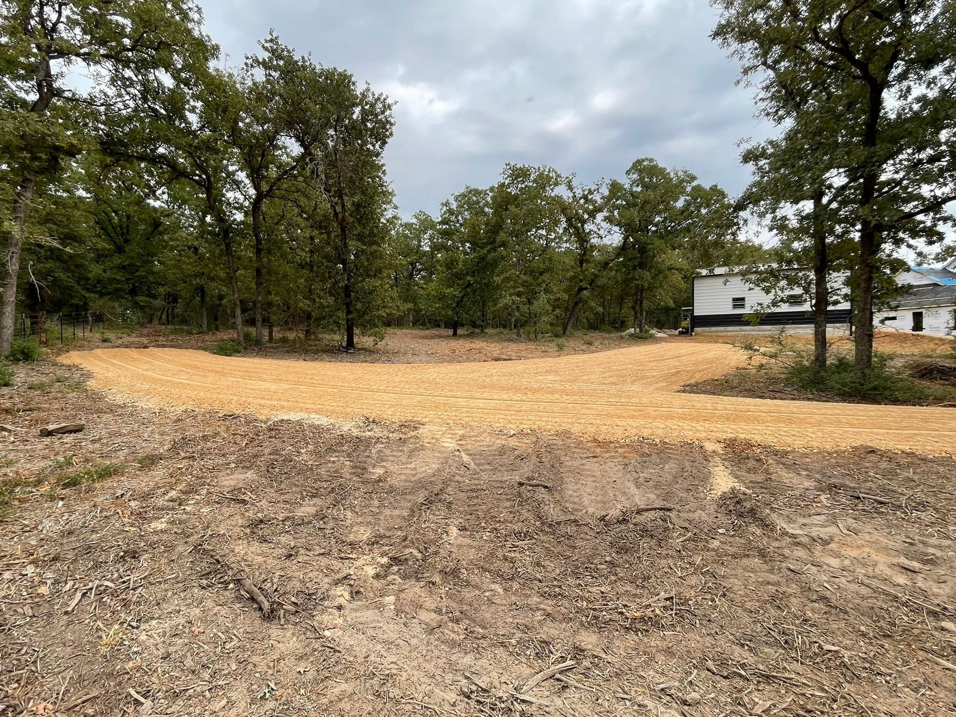 A gravel road curves through a wooded area with trees and a building in the background.