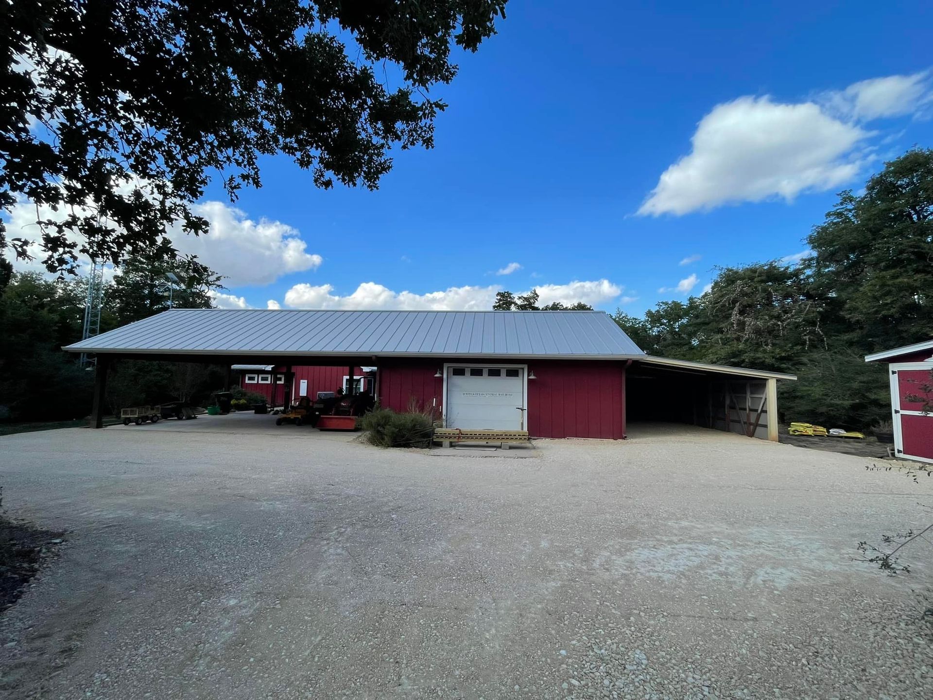 Red barn with carport under blue sky, gravel driveway.