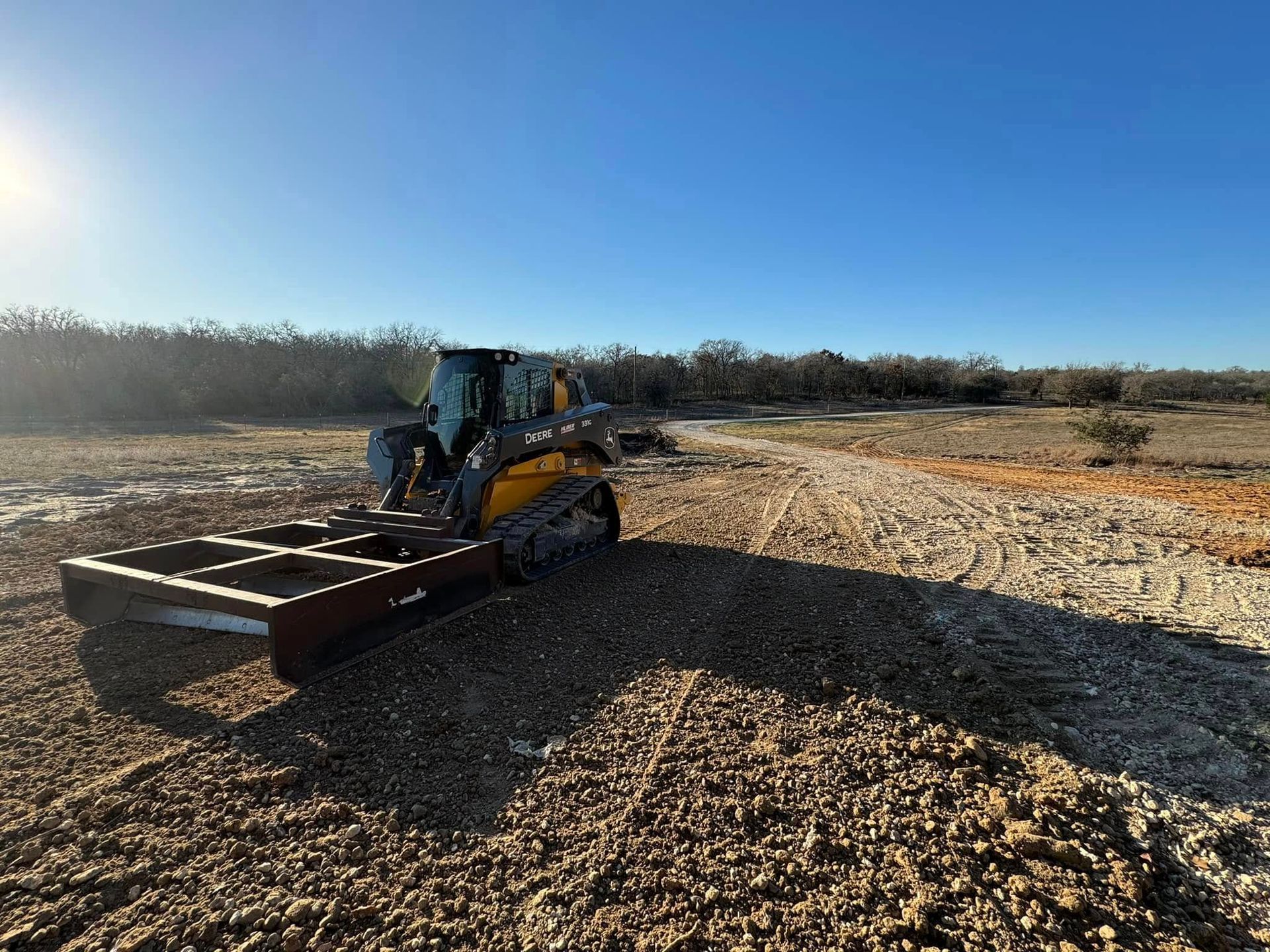 A track skid steer with a flat attachment grading a gravel path under a clear blue sky.
